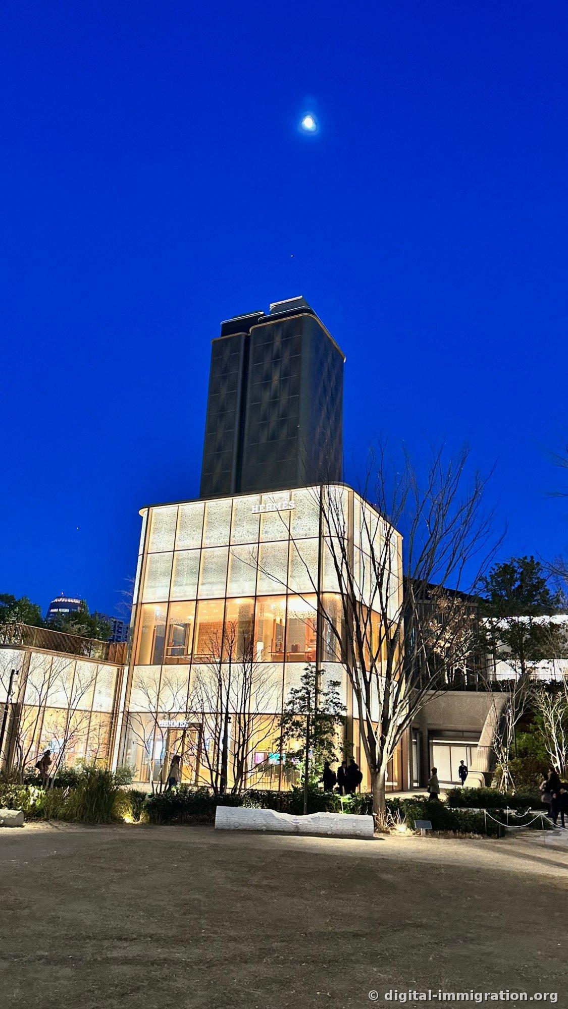 Looking up at Hermès lower level and dark high tower, with moon in deep blue sky and white bench in the foreground