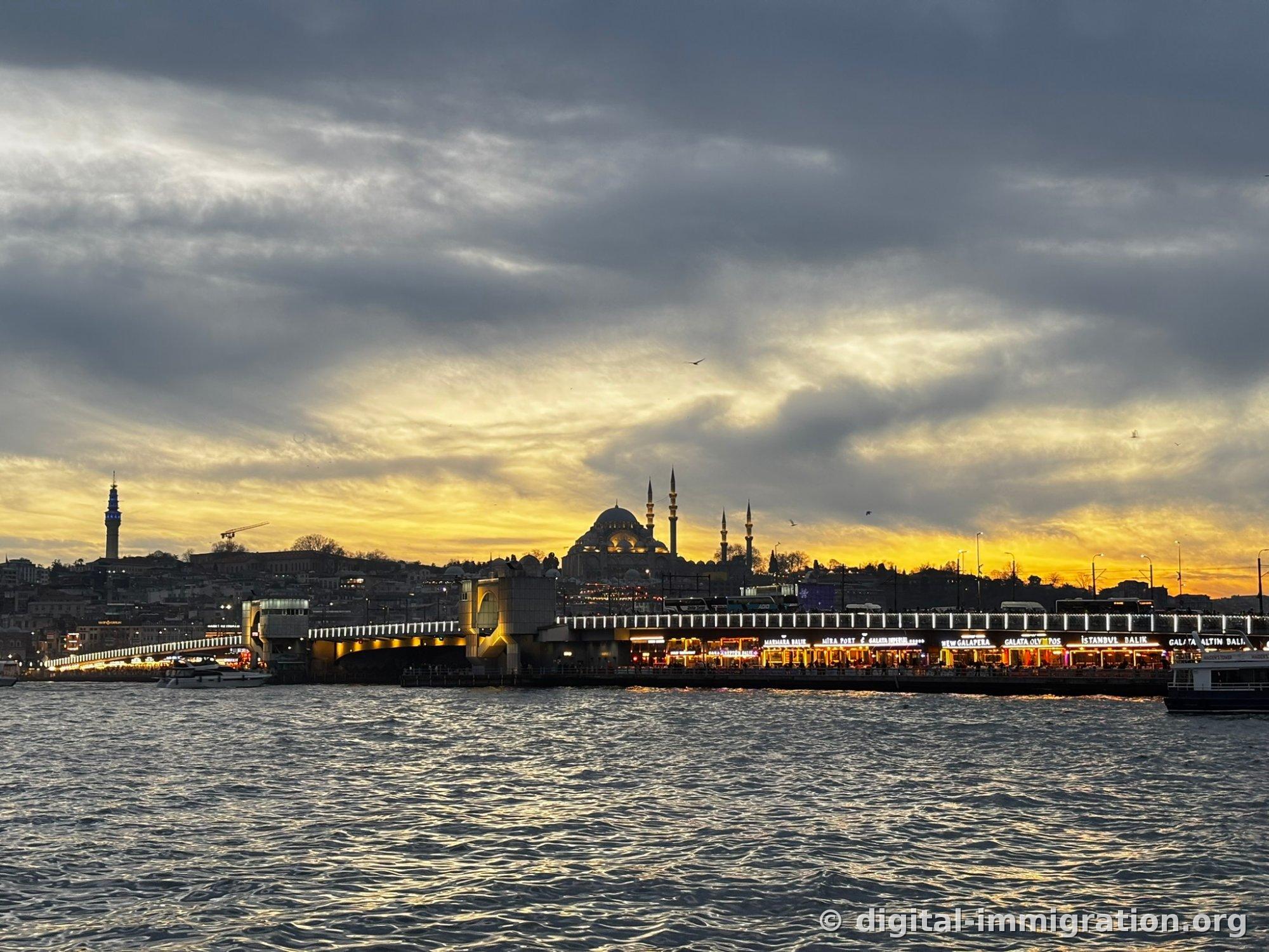 Yellow-tinged sky under thick clouds, Galata Bridge lights, fish restaurant signs on the lower deck of the bridge, silhouette of Süleymaniye Mosque on the hill above