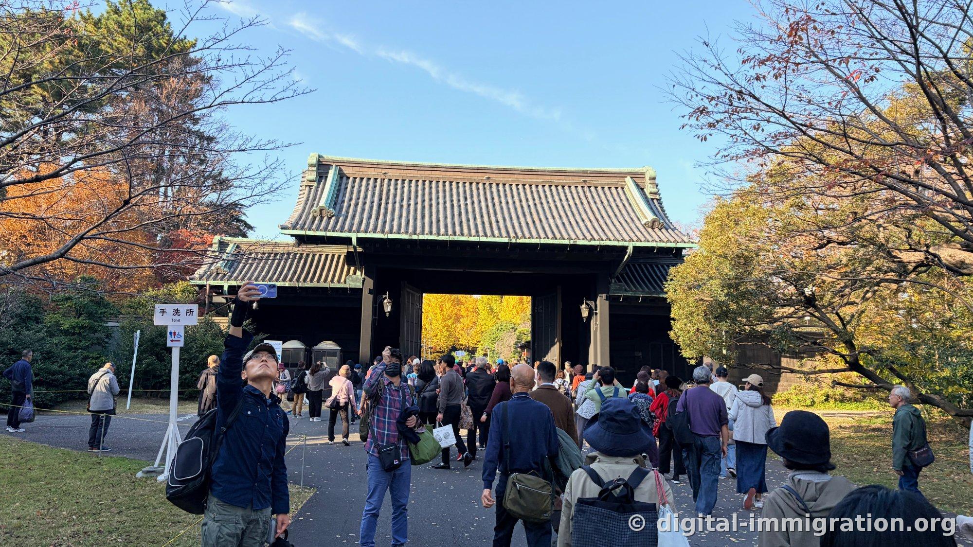 Un grand portail fortifié avec un ginkgo jaune cadré par l'arche, beaucoup de gens prenant des photos pour passer, avec un panneau de toilettes à gauche
