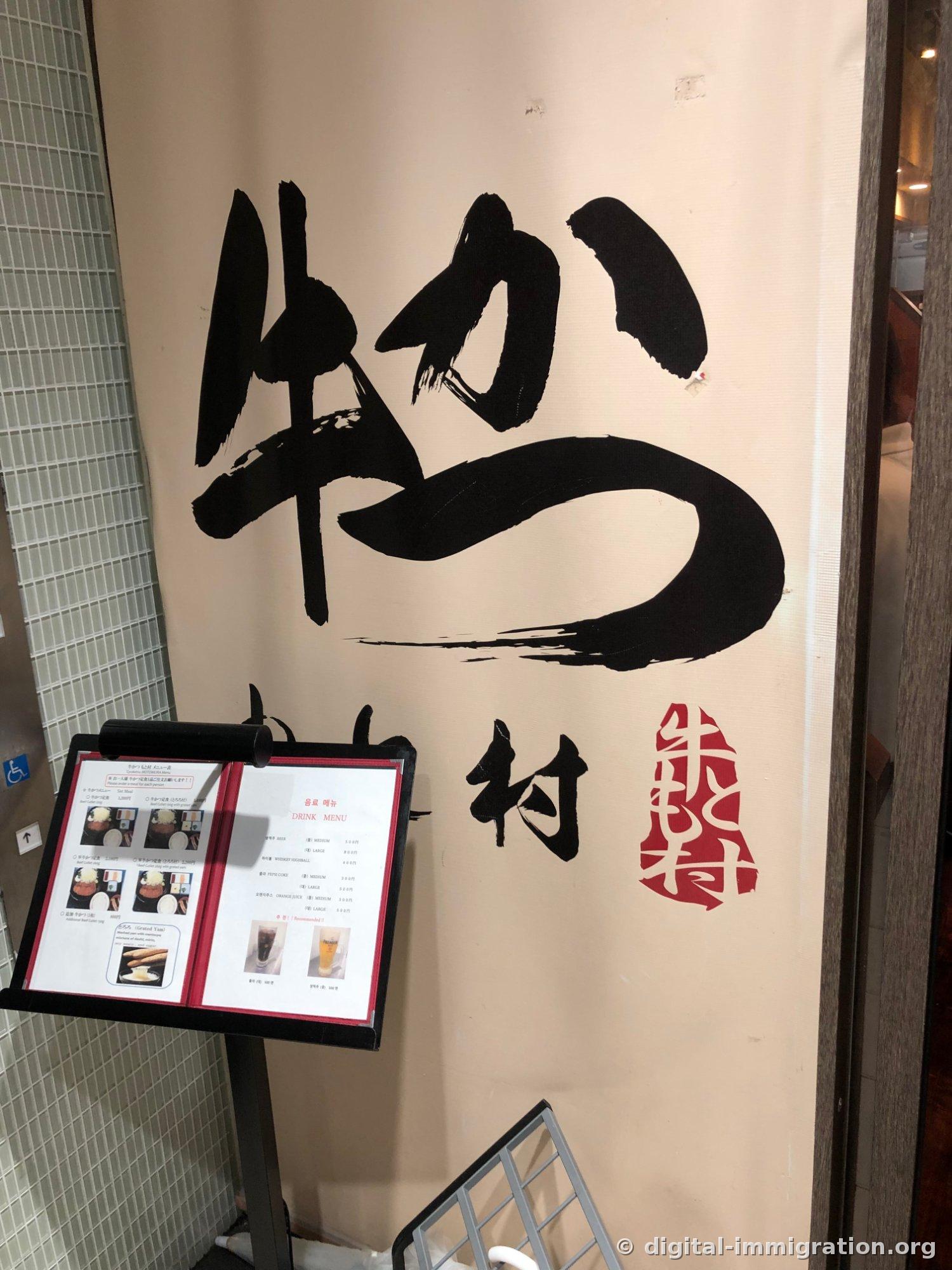 L'entrée de Gyukatsu Motomura avec le noren crème écrit à la main 牛かつ もと村 et un cachet rouge, devant un présentoir de menu avec photos