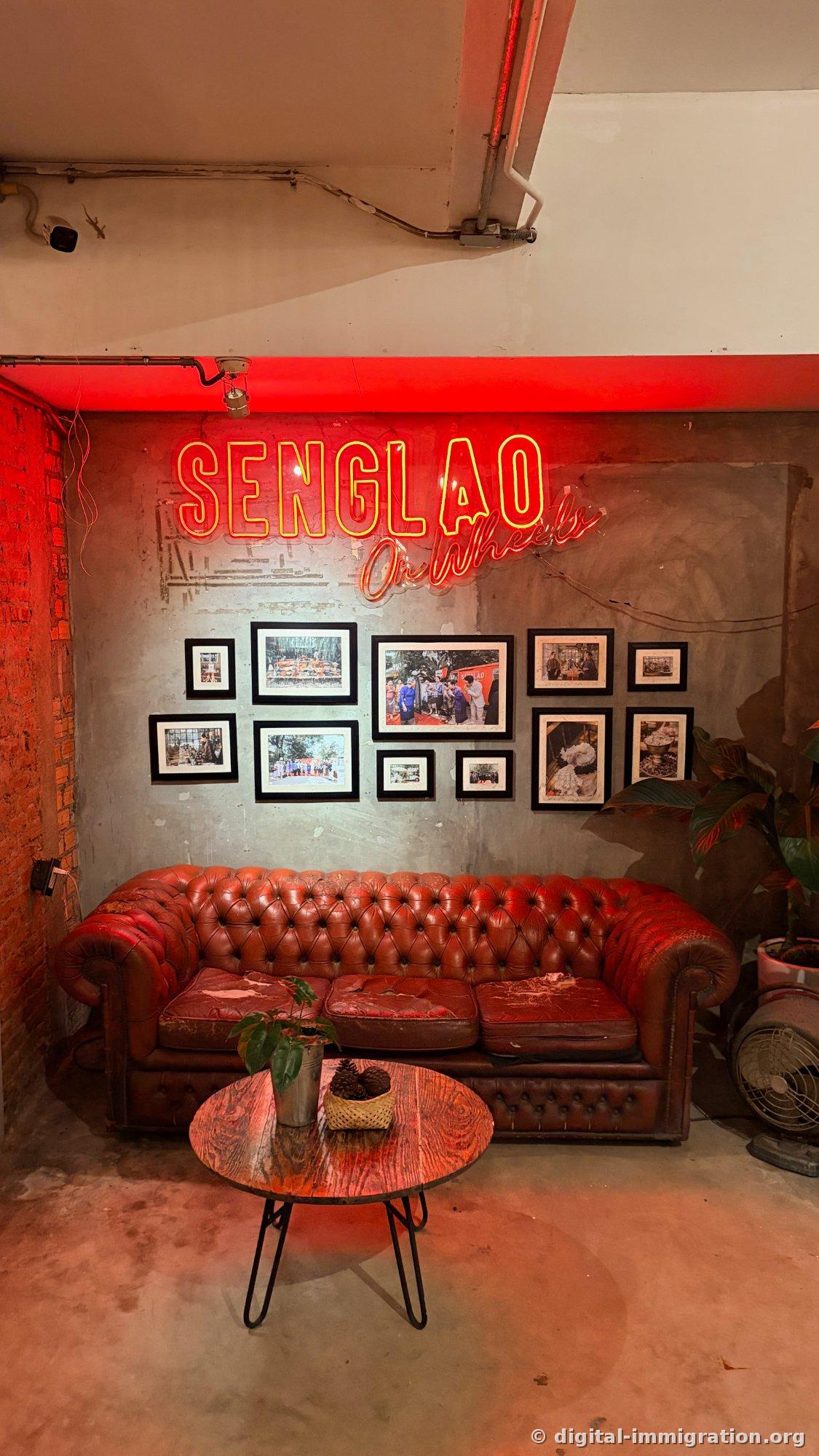 Un néon rouge 'SENGLAO On Wheels' au-dessus d'un mur de ciment avec neuf photos encadrées et un vieux canapé Chesterfield en cuir rouge usé, une petite table ronde en bois