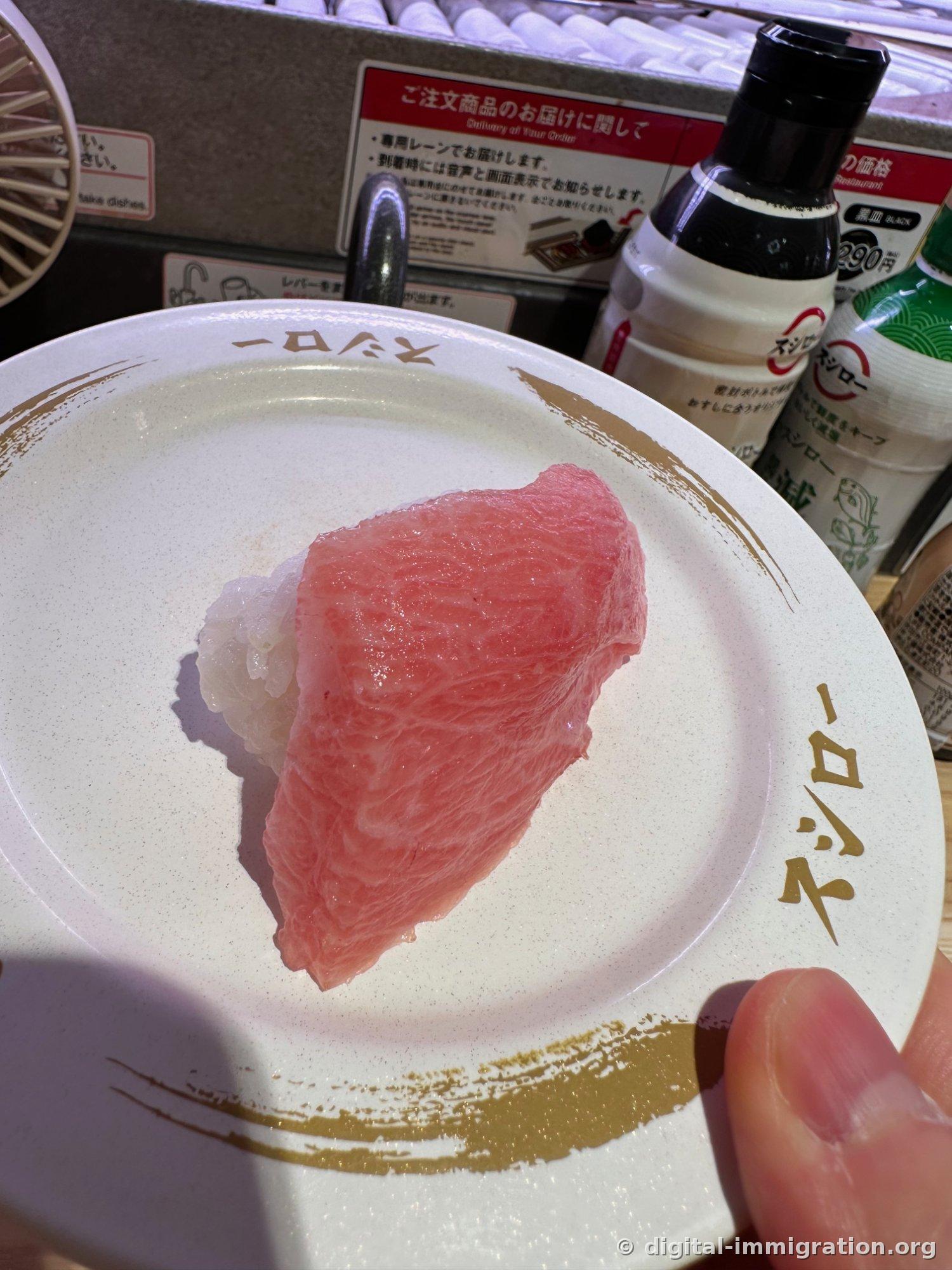 금빛 무늬가 그려진 흰 스시로 접시 위에 놓인 두툼한 참치 대뱃살 초밥. 뒤로 간장병과 저염 간장병이 보인다