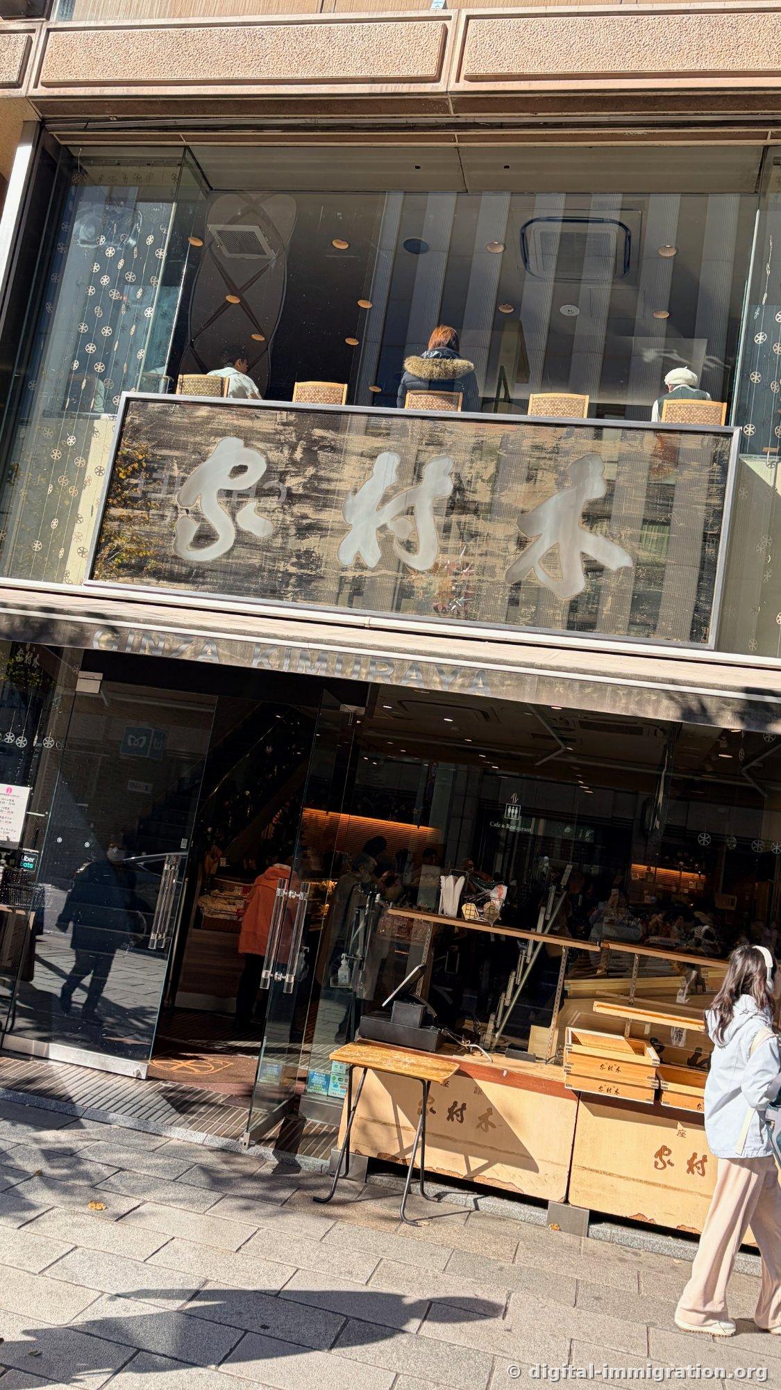 Winter exterior of Kimuraya with sunlit cursive wooden signboard, 'GINZA KIMURAYA' text and stacked wooden box outdoor stand visible