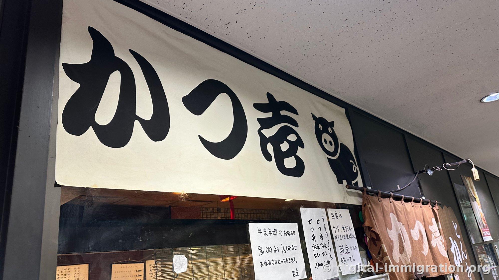 The shop's awning sign with 'かつ庵' written in black brushstrokes on a white background and a drawing of a pig