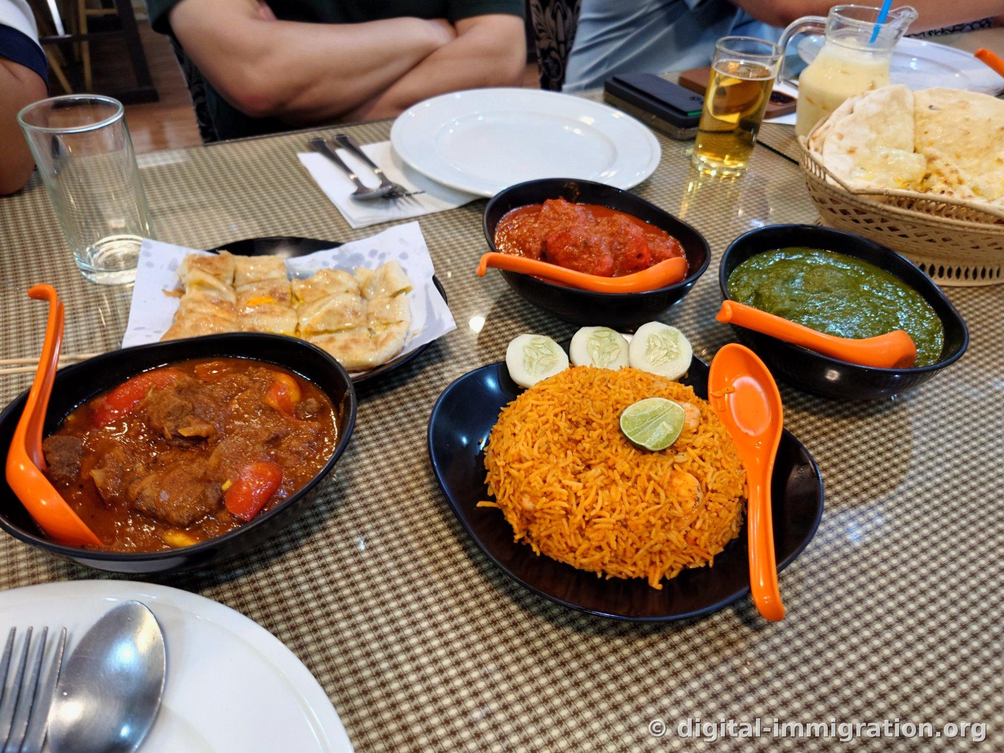 The table laden with red mutton curry, tandoori chicken, green palak, shrimp biryani, and a basket of naan, roti, and glasses of Beerlao