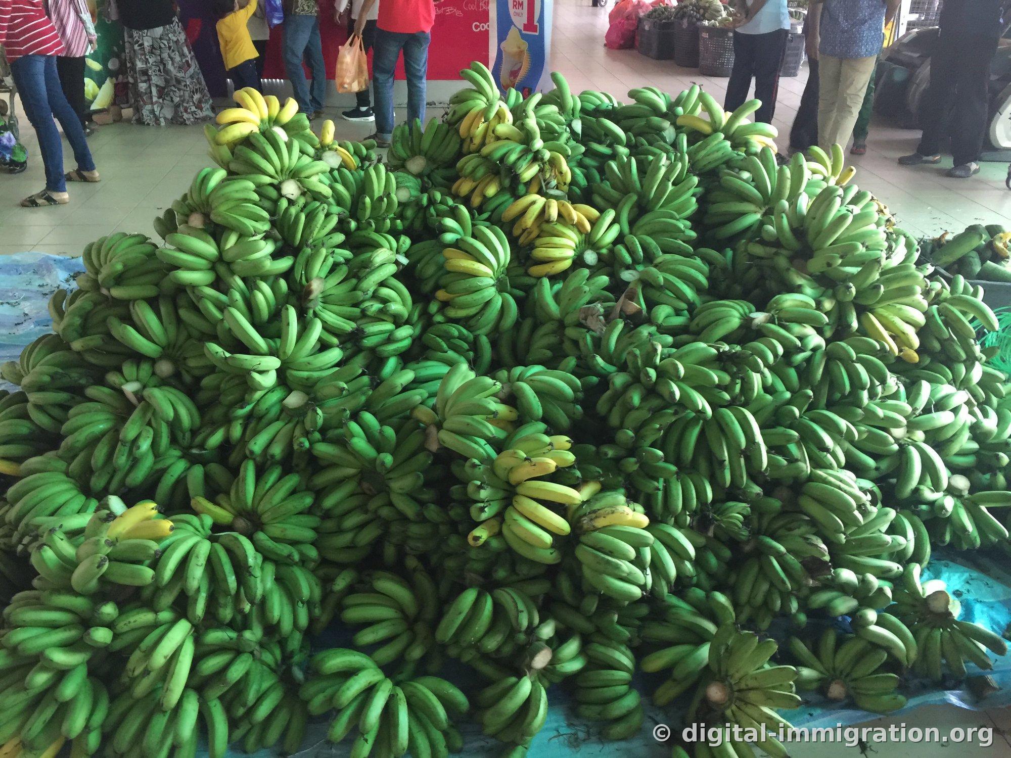 Des régimes de bananes vertes empilés comme une montagne sur une bâche bleue posée sur le sol du marché, avec les jambes des clients allant et venant autour, et une banneau publicitaire pour la crème glacée rouge visible à l'arrière