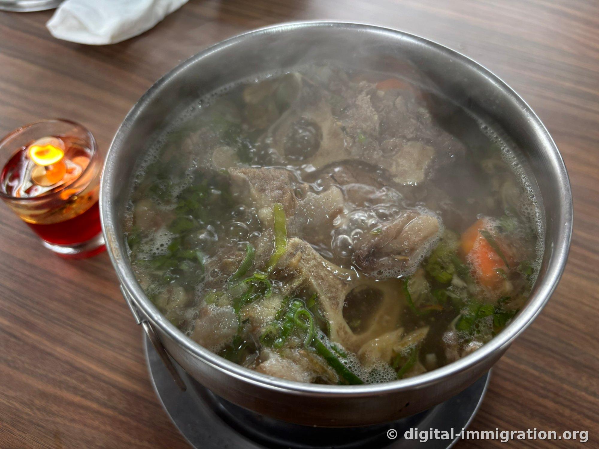 Clear oxtail soup simmering in a stainless steel pot over solid fuel, with bone and scallion and carrot visible, and a container of burning fuel tablet to the side