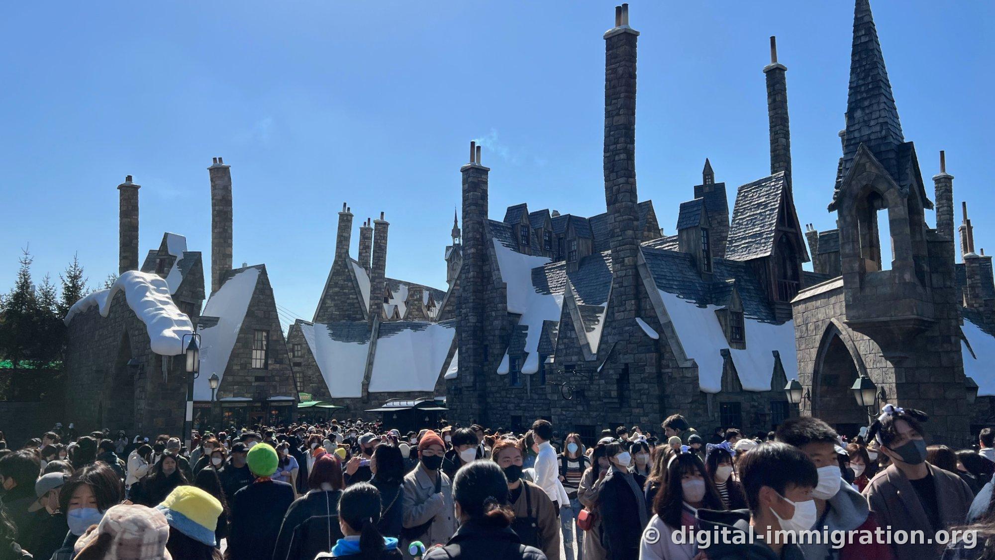 Panoramic view of Hogsmeade village with thickly snow-covered gray roofs and brick chimneys of varying heights, one chimney emitting smoke, with the lane below crowded with masked visitors