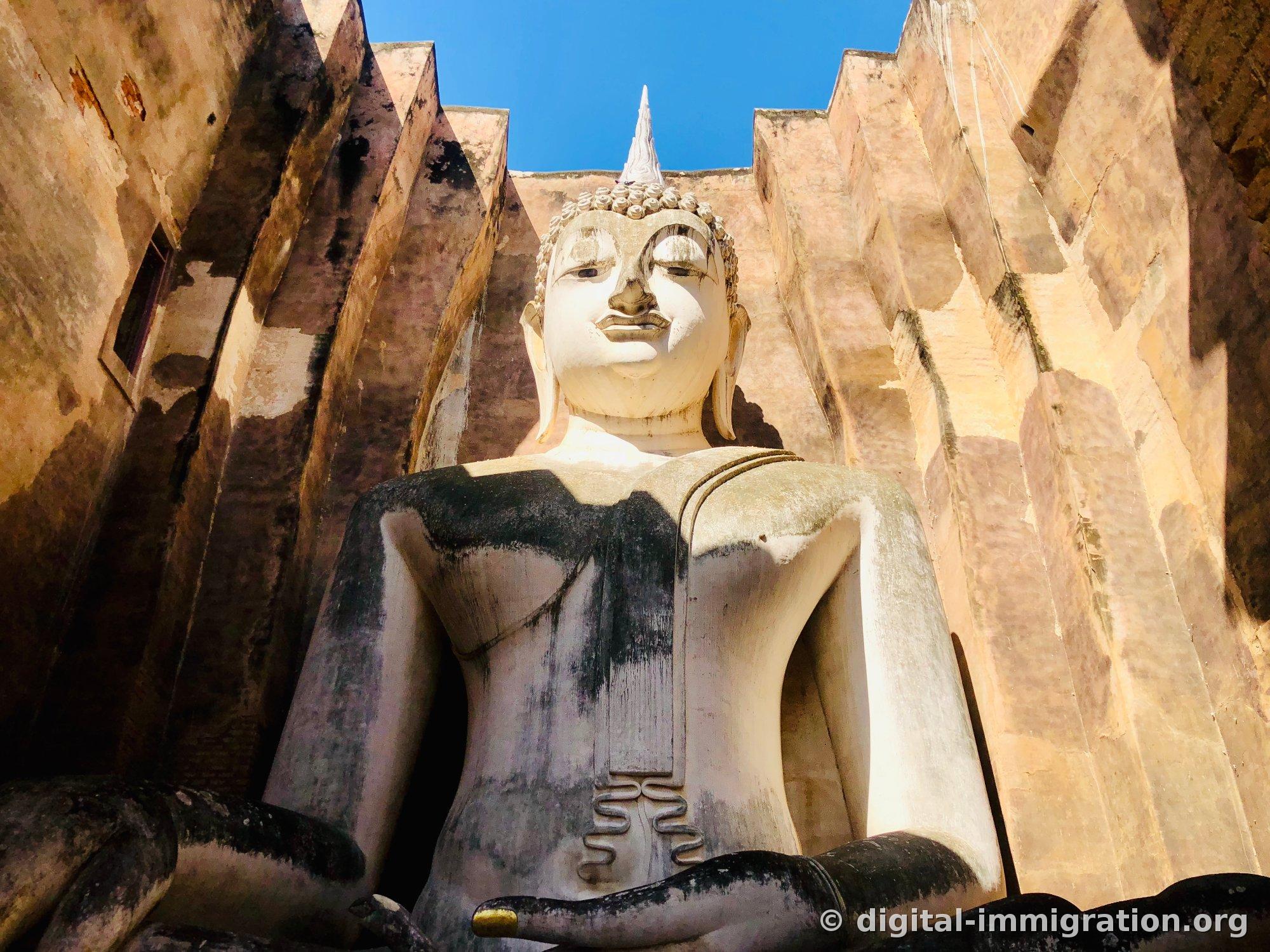 Phra Achana, a seated Buddha looking up from inside Wat Si Chum, with a white face and a darkly stained body, with gold leaf visible on the fingertips