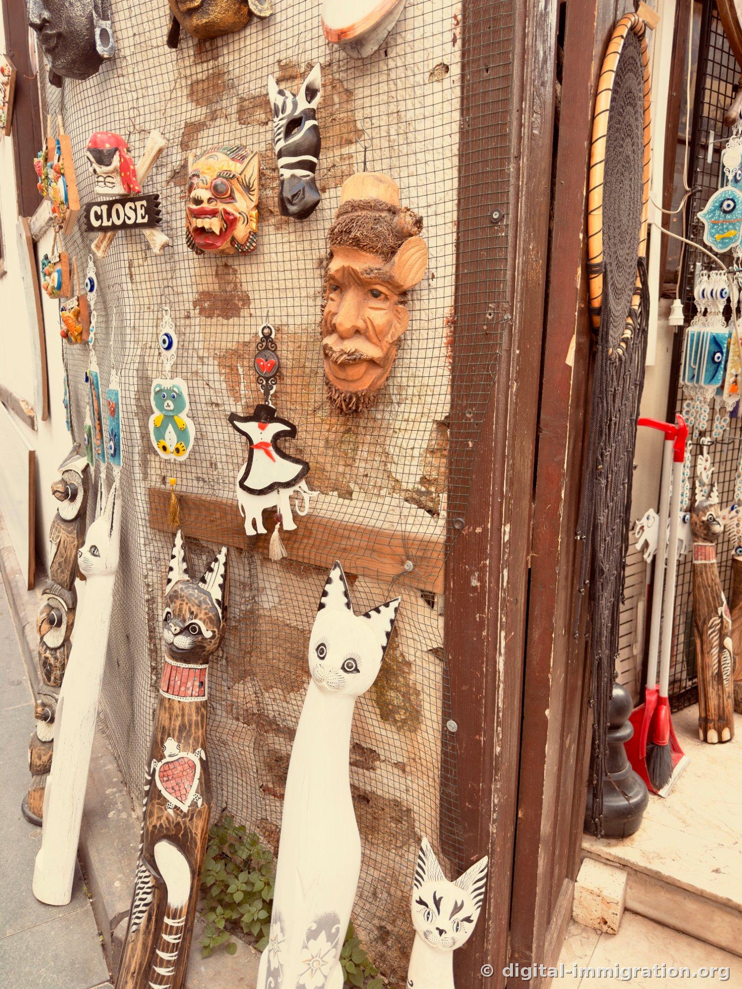 Façade d'une boutique de souvenirs avec des masques en bois et des décorations bleues de nazar boncuk accrochées au treillis, et des sculptures de chats en bois blanc et brun debout au sol. Au centre, une plaque en bois avec le mot 'CLOSE'