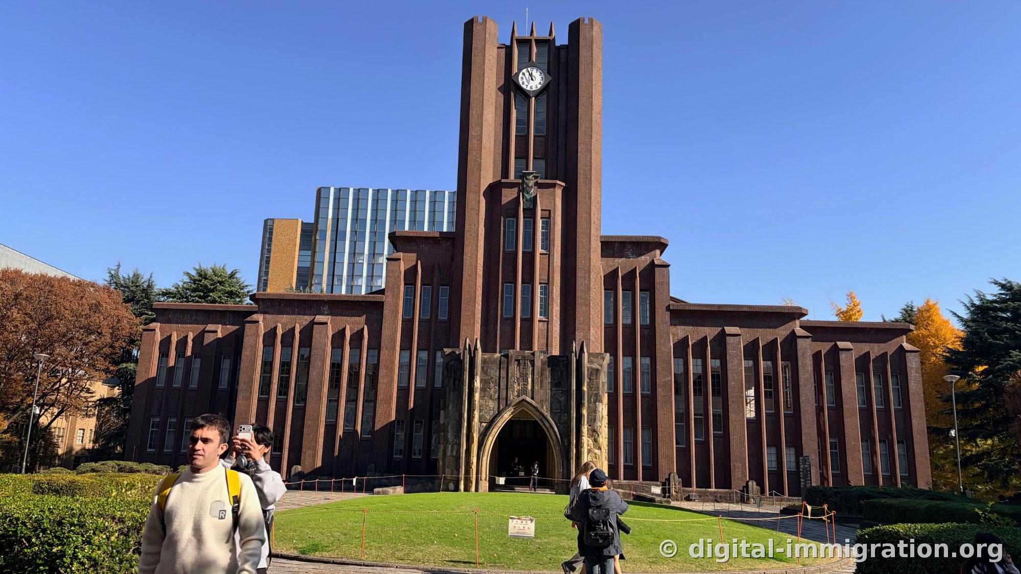Red brick facade of Tokyo University's Yasuda Auditorium under blue sky, with a clock tower in the center, a pointed arched entrance, and a circular lawn in front