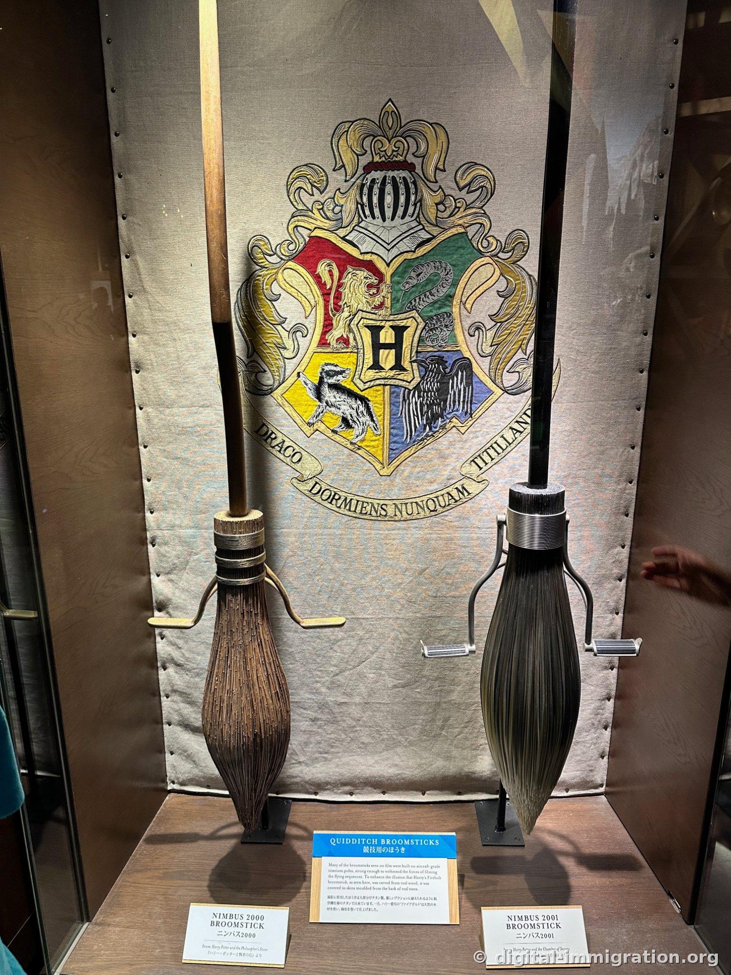 Two broomsticks standing in a glass case against a backdrop showing the Hogwarts crest