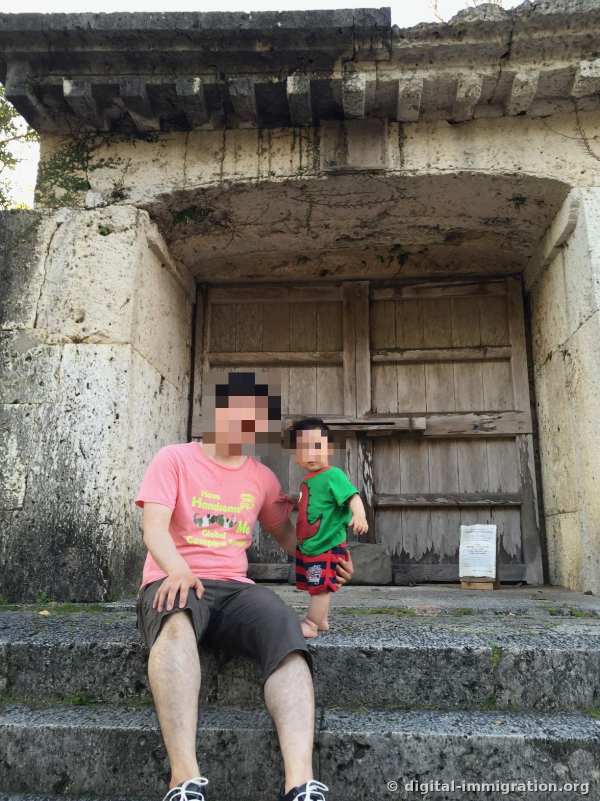 An adult and child sitting on stone steps in front of the weathered wooden gate of Sonohouguu Utaki Stone Gate built from Ryukyu limestone