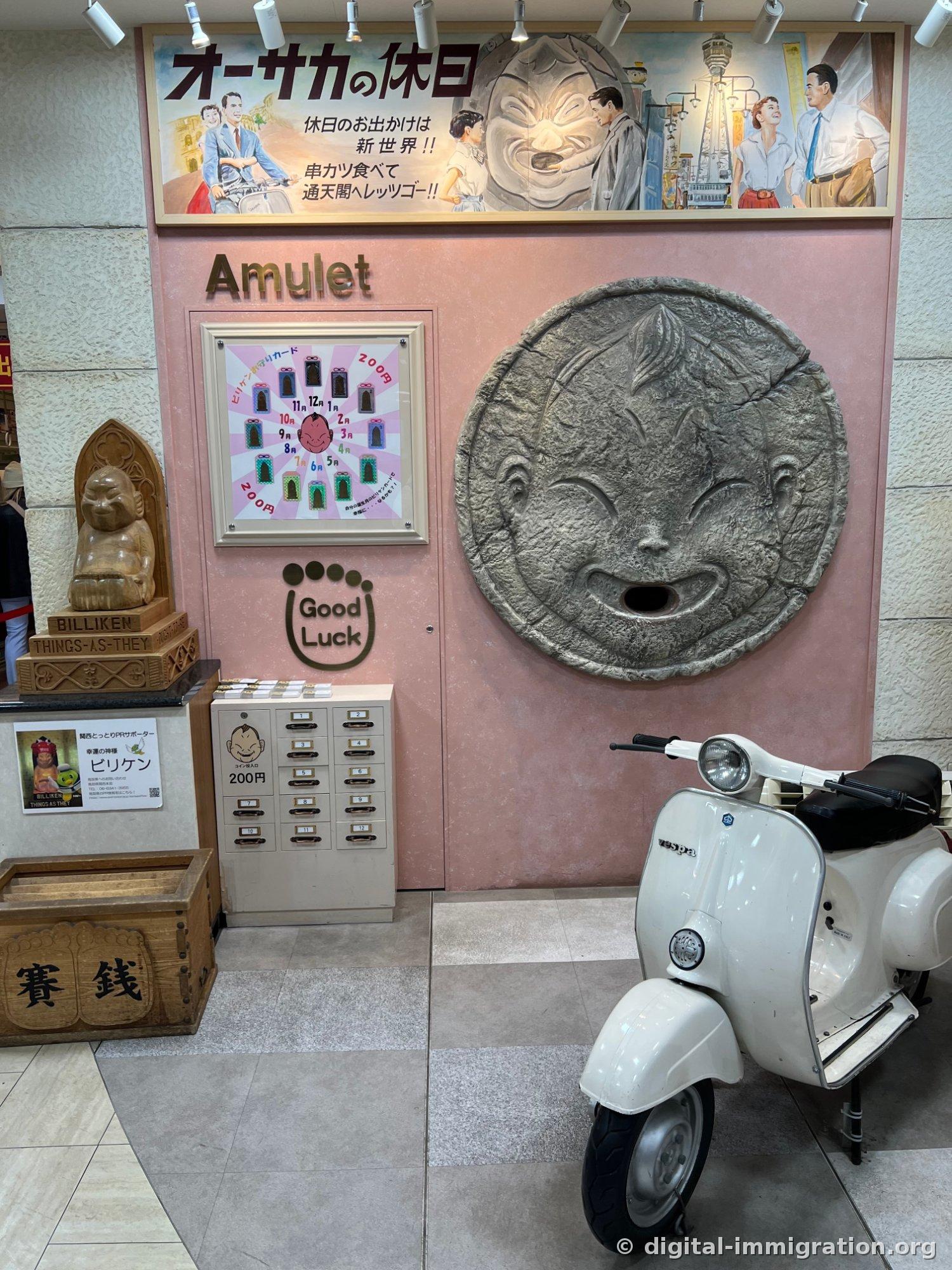 An overall view of the Billiken corner, with a wooden Billiken statue and coin purse on the left, a 200-yen charm-dispensing drawer in the center