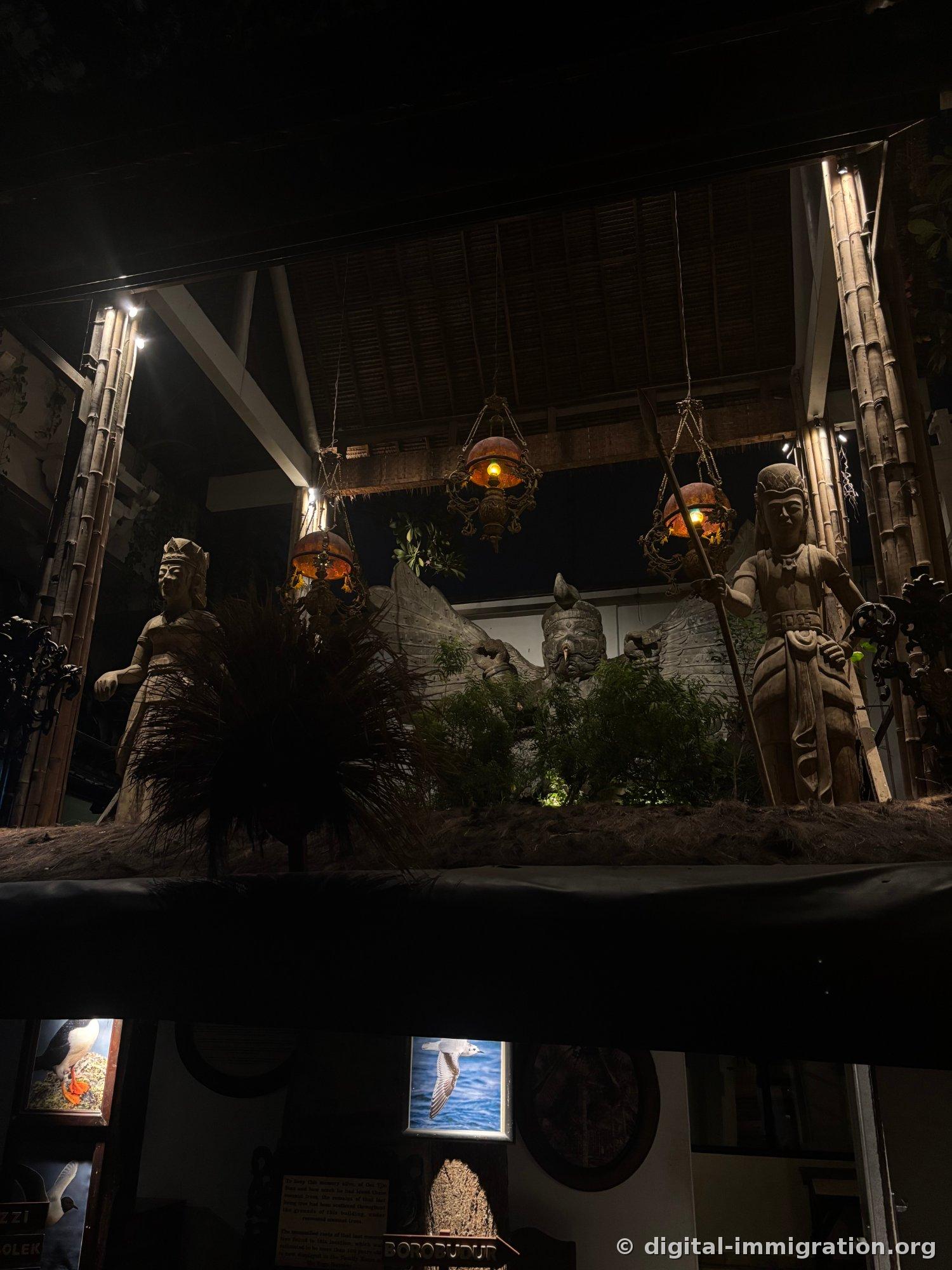 Dark exhibition space under bamboo posts and thatched roof with brass lanterns hanging and stone statues holding windows facing each other, BOROBUDUR sign below