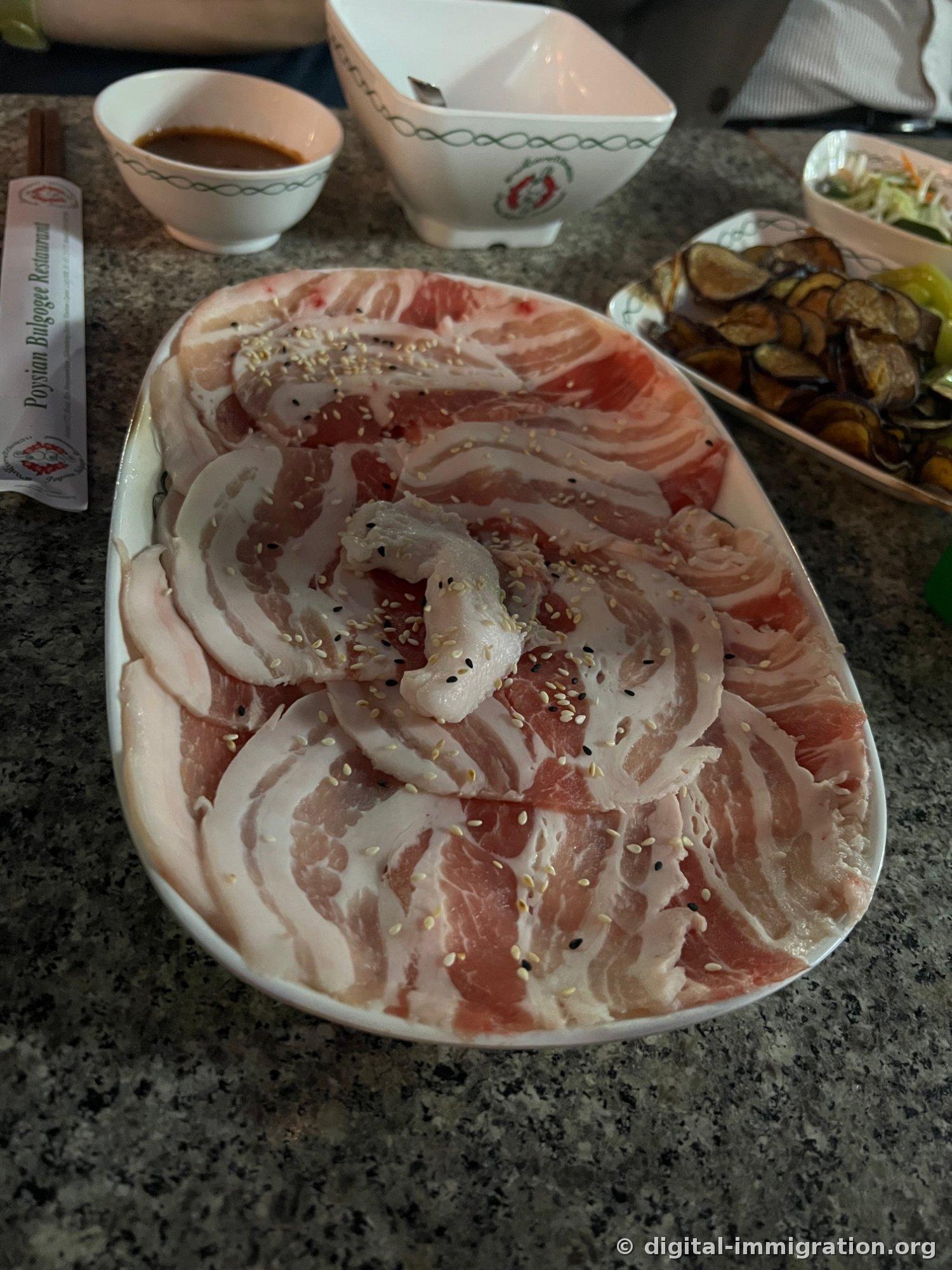 Raw pork belly rolled like a rose and sprinkled with sesame on a white plate, with a bowl of brown sauce beside it, and chopstick wrapper labeled 'Poysian Bulgogee Restaurant'
