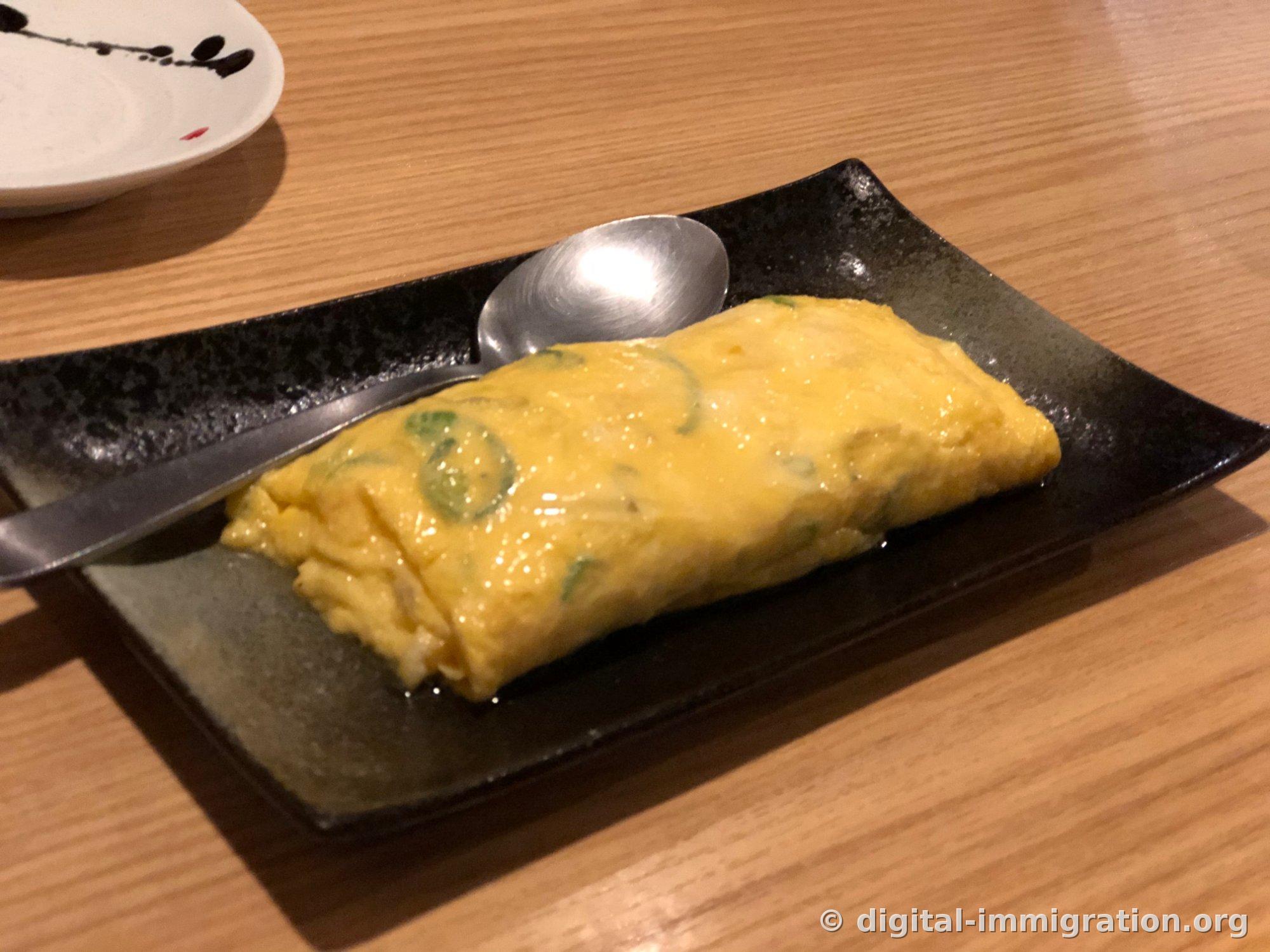 검은 직사각 접시에 놓인 통통한 다시마키 계란말이와 은색 스푼