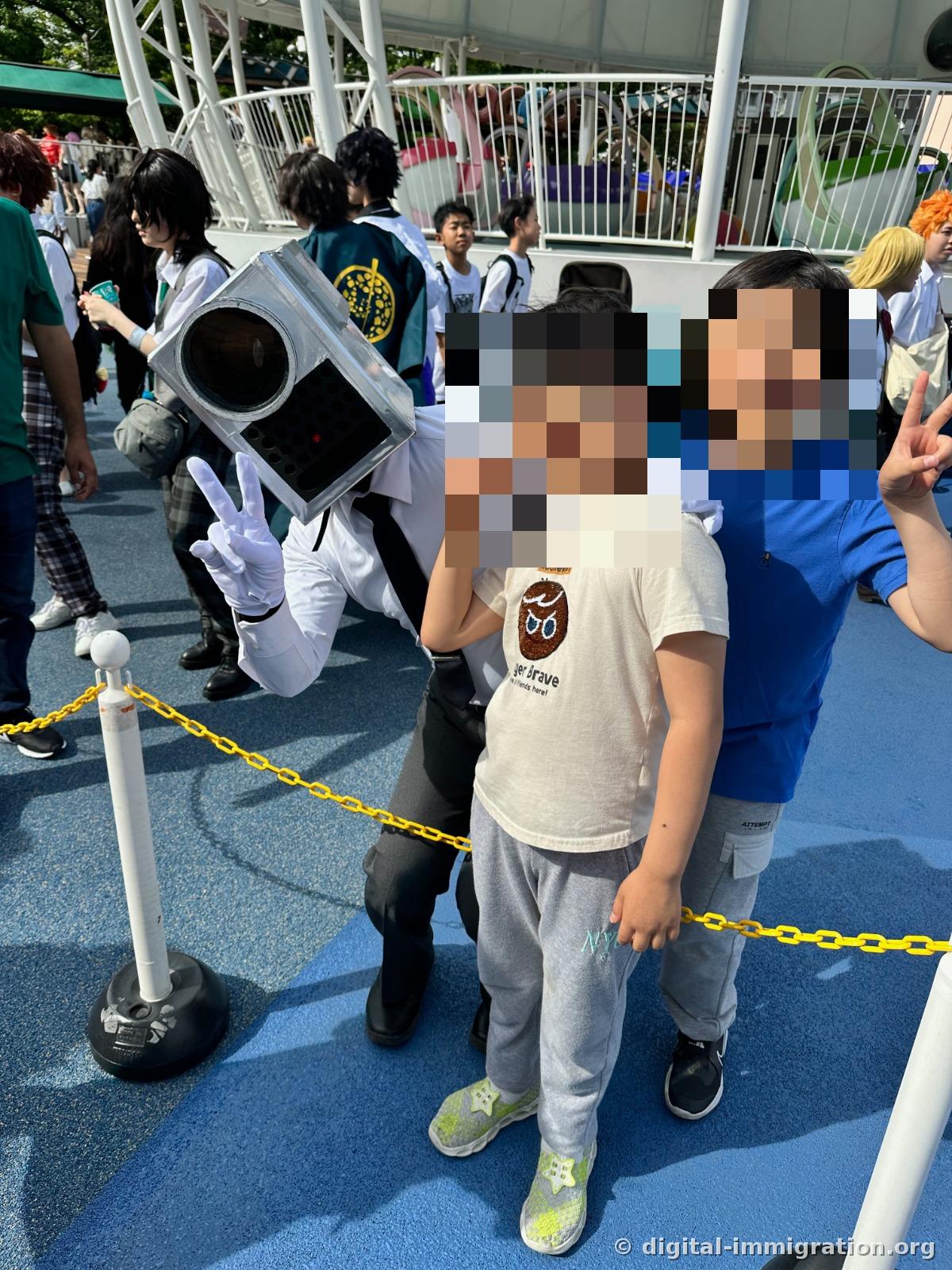 Dans la zone de chaîne jaune du plancher bleu du parc d'attractions, un participant en cosplay portant une boîte en forme de caméra argentée sur la tête et posant un V avec des gants blancs, avec deux enfants posant ensemble devant lui