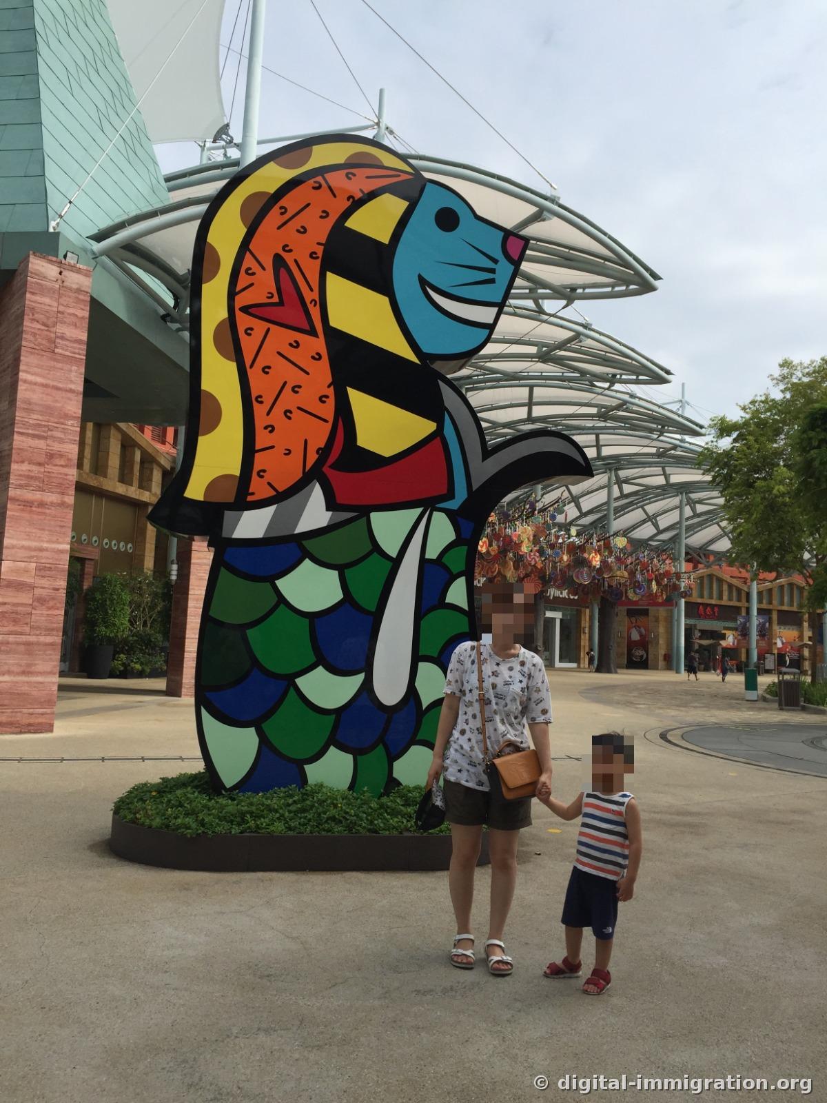 An adult and child holding hands in front of a pop-art Merlion sculpture painted with primary color blocks and fish scale patterns