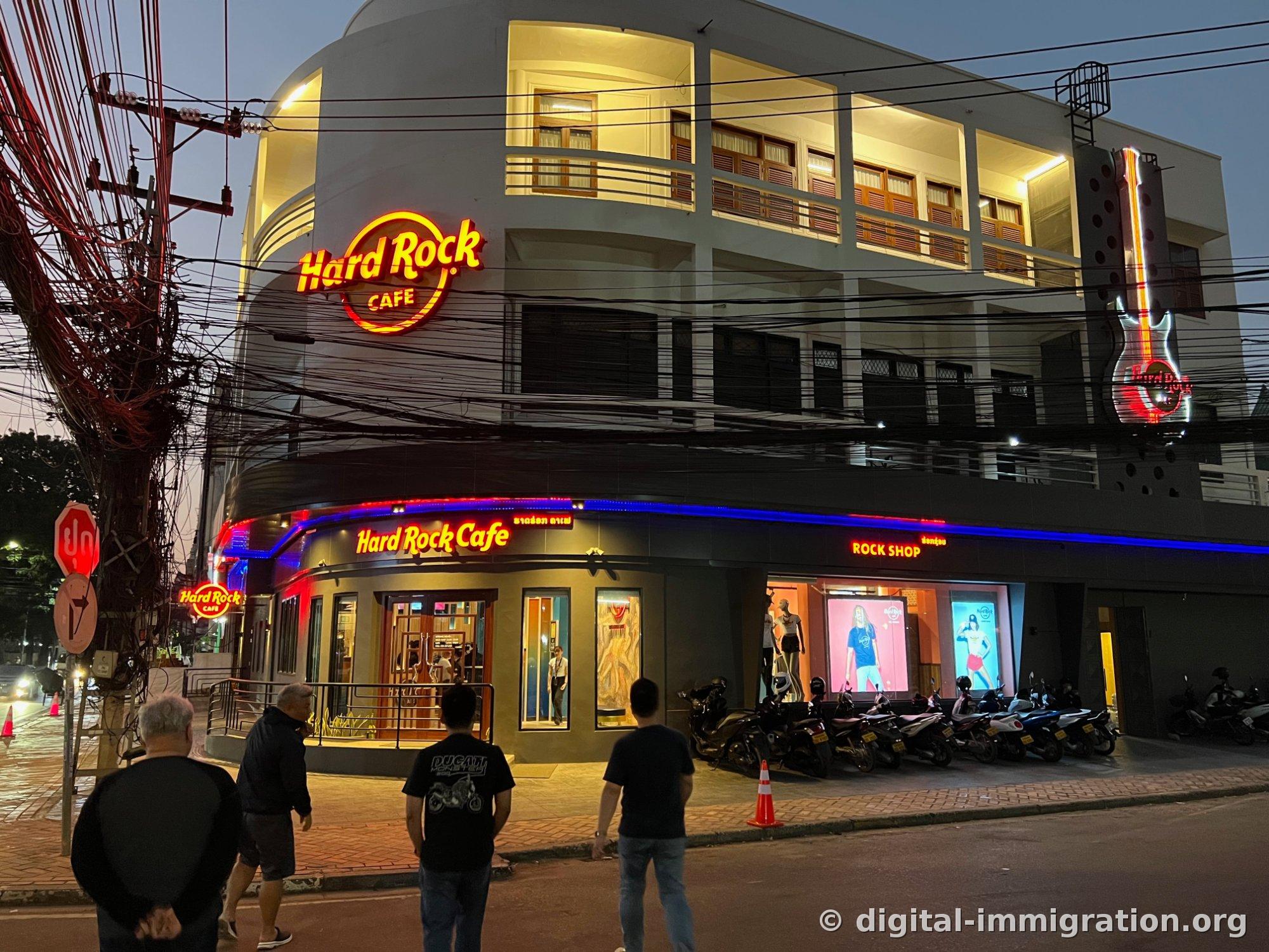 어둑해진 저녁, 곡선 코너 건물에 붉은 Hard Rock Cafe 원형 네온과 파란 띠 조명이 켜져 있고 앞쪽에 오토바이가 줄지어 주차된 전경