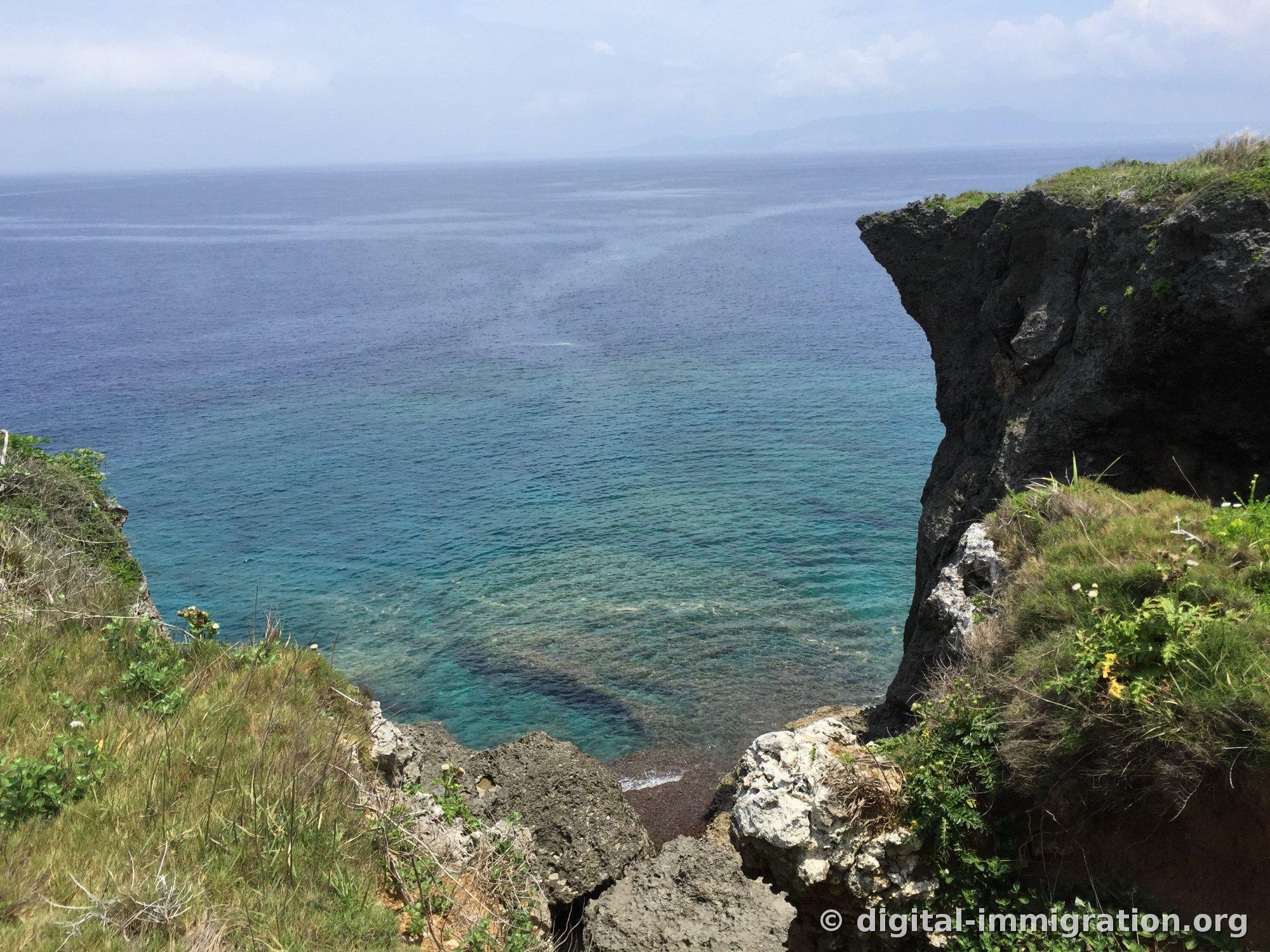 草で覆われた崖の間から見下ろした万座毛の湾。右側に黒い石灰岩の崖が突き出ており、水面下にはサンゴのパターンが見えている