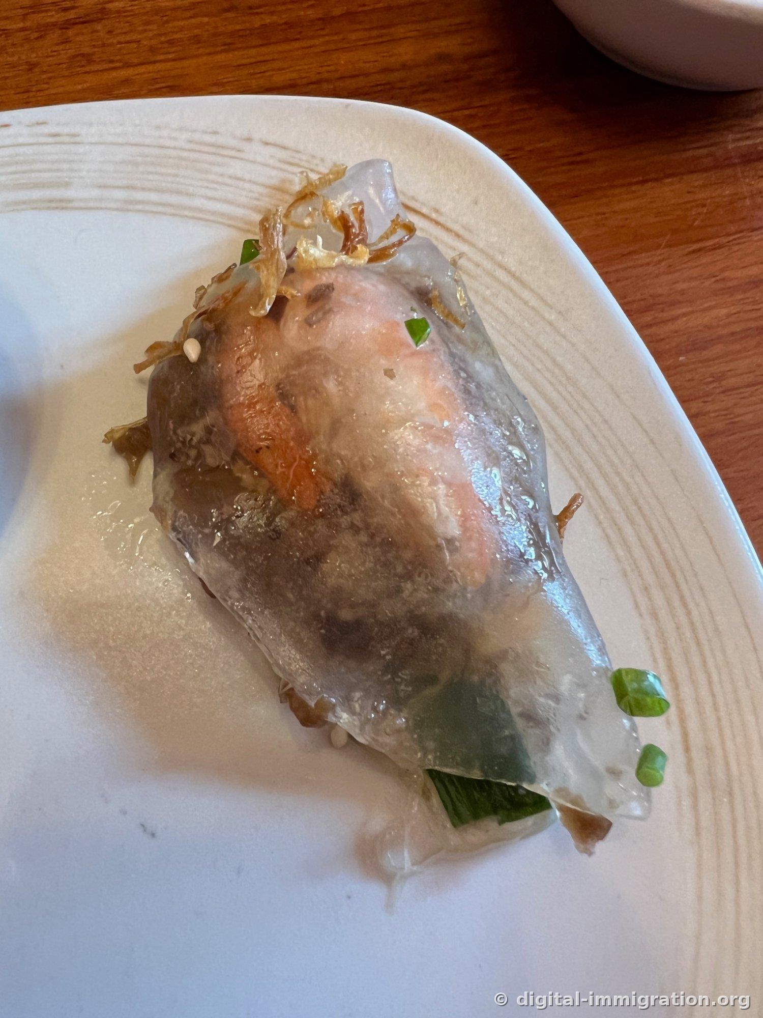 접시에 덜어놓은 라이스페이퍼 롤 한 개를 가까이 찍은 사진, 반투명한 피 사이로 새우와 버섯, 쪽파가 비쳐 보이고 위에 튀긴 샬롯이 얹혀 있다