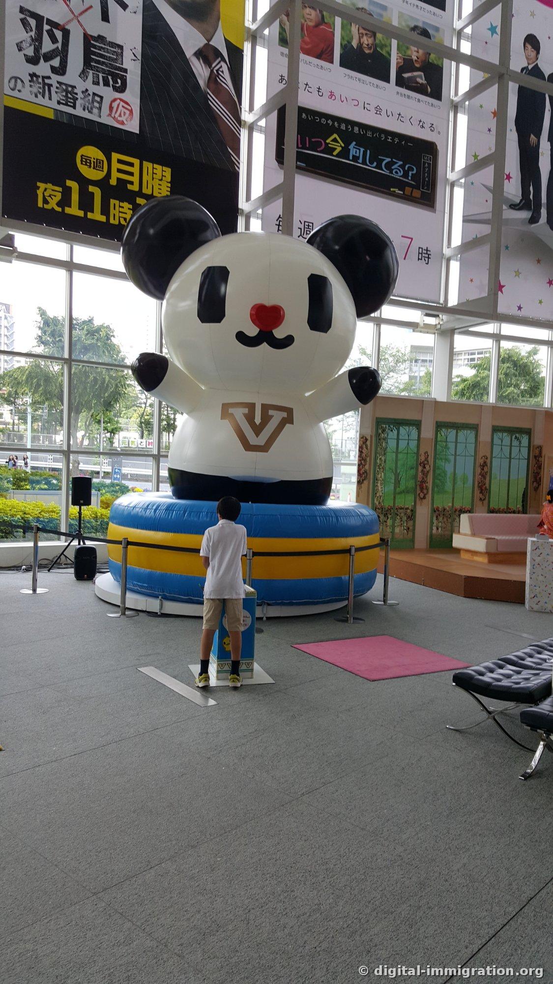 Un gigantesque personnage de panda gonflable dressé à l'intérieur d'un atrium aux murs vitrés avec un enfant en t-shirt blanc debout devant et levant les yeux vers lui, des bannières de programmes de télévision accrochées au plafond