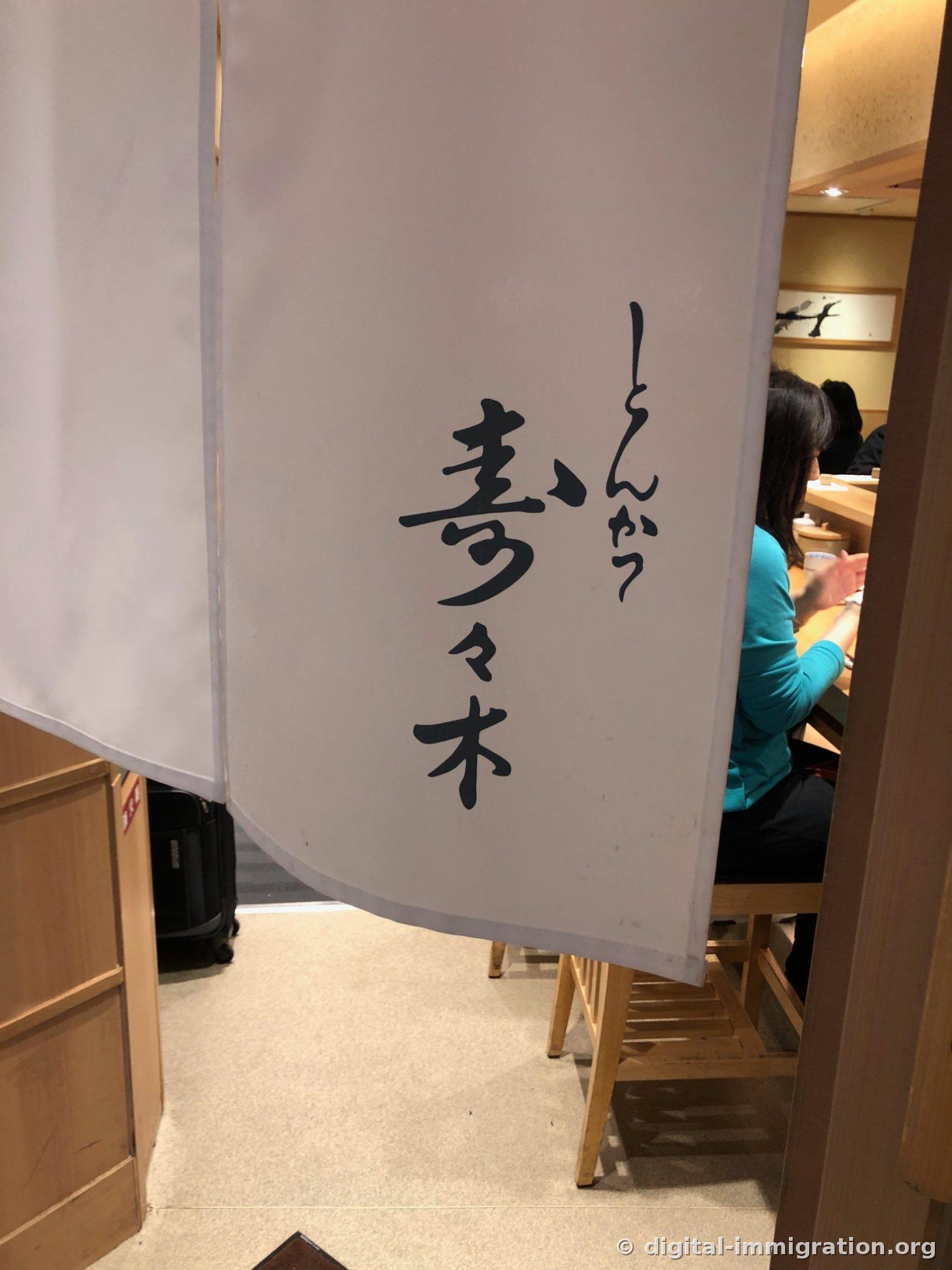 白色暖帘上用黑笔写着'とんかつ 寿々木'，缝隙里能看到坐在吧台的顾客和行李箱