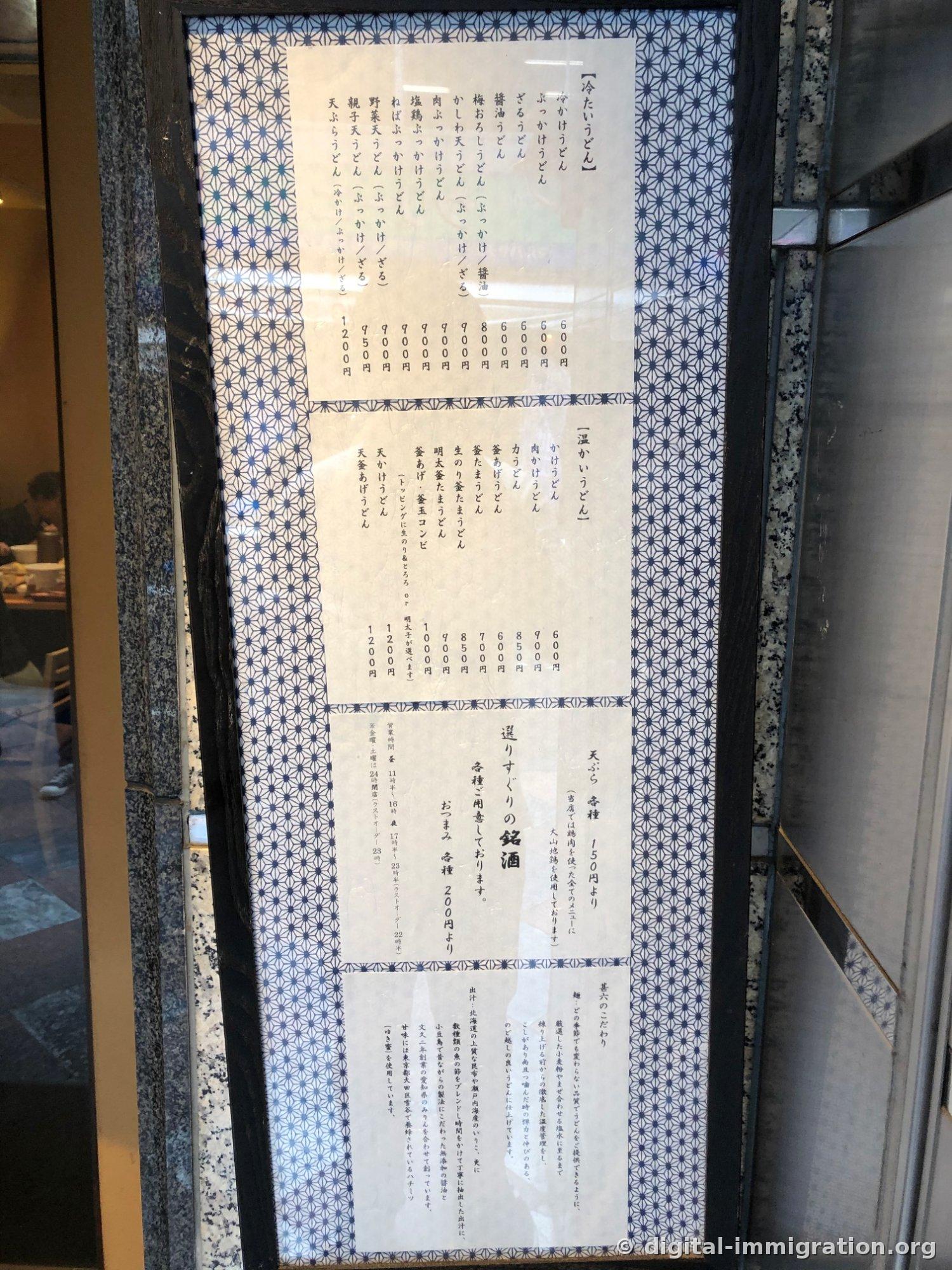 店のドア横のガラス額に貼られた日本語縦書きメニュー板、冷うどんと温うどんの価格が600円から1200円まで書かれている