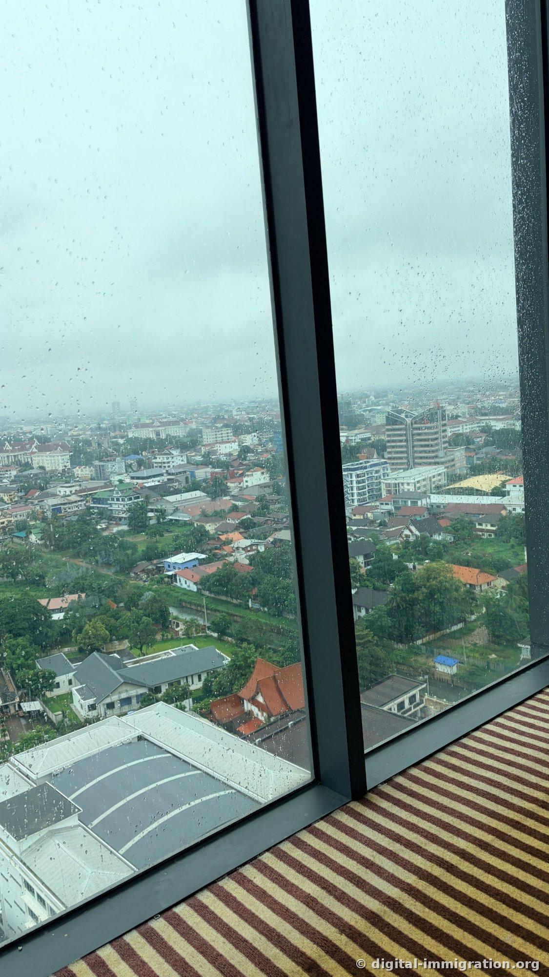 Vue depuis la fenêtre en verre de la chambre d'étage élevé avec des gouttes de pluie, montrant la ville de Vientiane sous un ciel nuageux et le tapis à rayures de la chambre