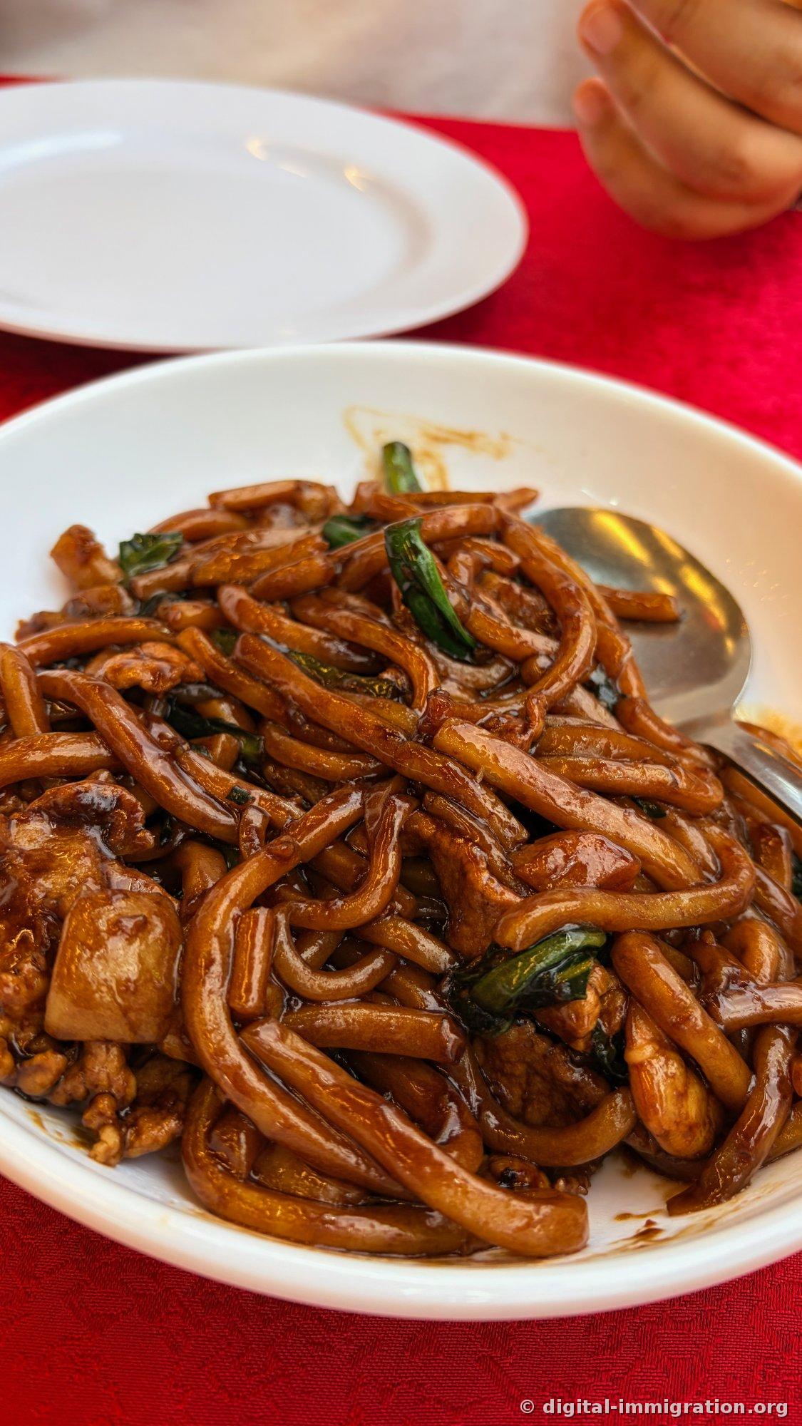 濃い茶色のソースがかかった太い麺にネギと肉が混ざったホッキエン麺が白いお皿に盛られ、赤いテーブルクロスの上に置かれている