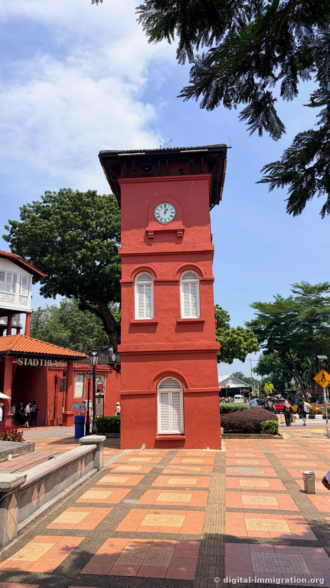The red brick clock tower and the STADTHUYS corridor on its left. The floor has orange checkered tiles, and a large tree behind creates shade
