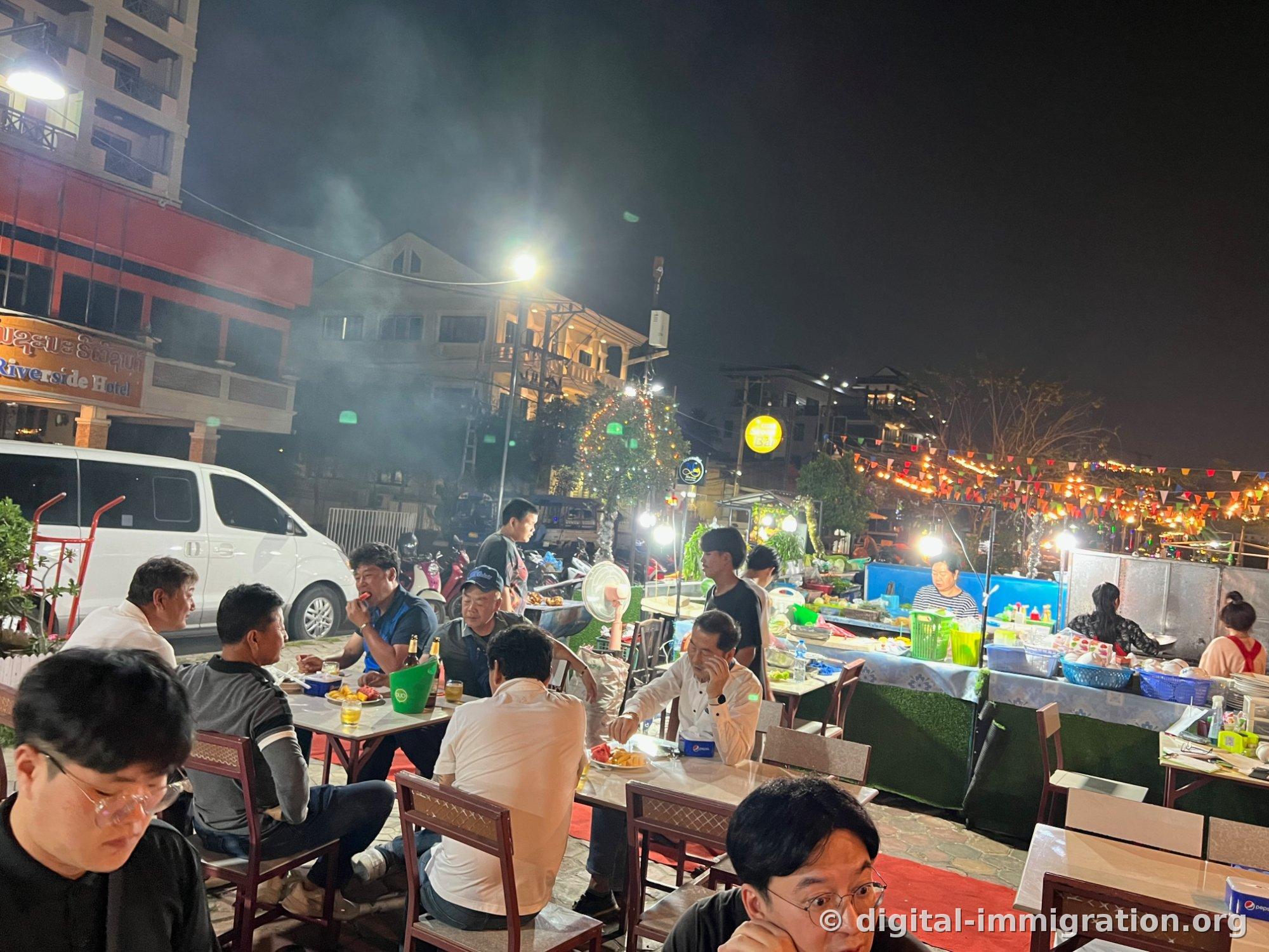 晚上靠路一侧的露天餐桌上人们在用餐，炭火烟气在路灯光下弥漫，背后能看到Riverside Hotel招牌和万国旗灯饰