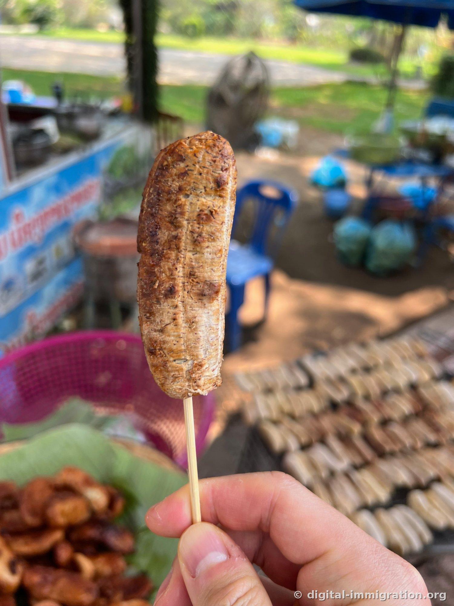 Une banane grillée marron tenue à la main sur une brochette, avec un grill en arrière-plan avec plusieurs bananes grillées alignées