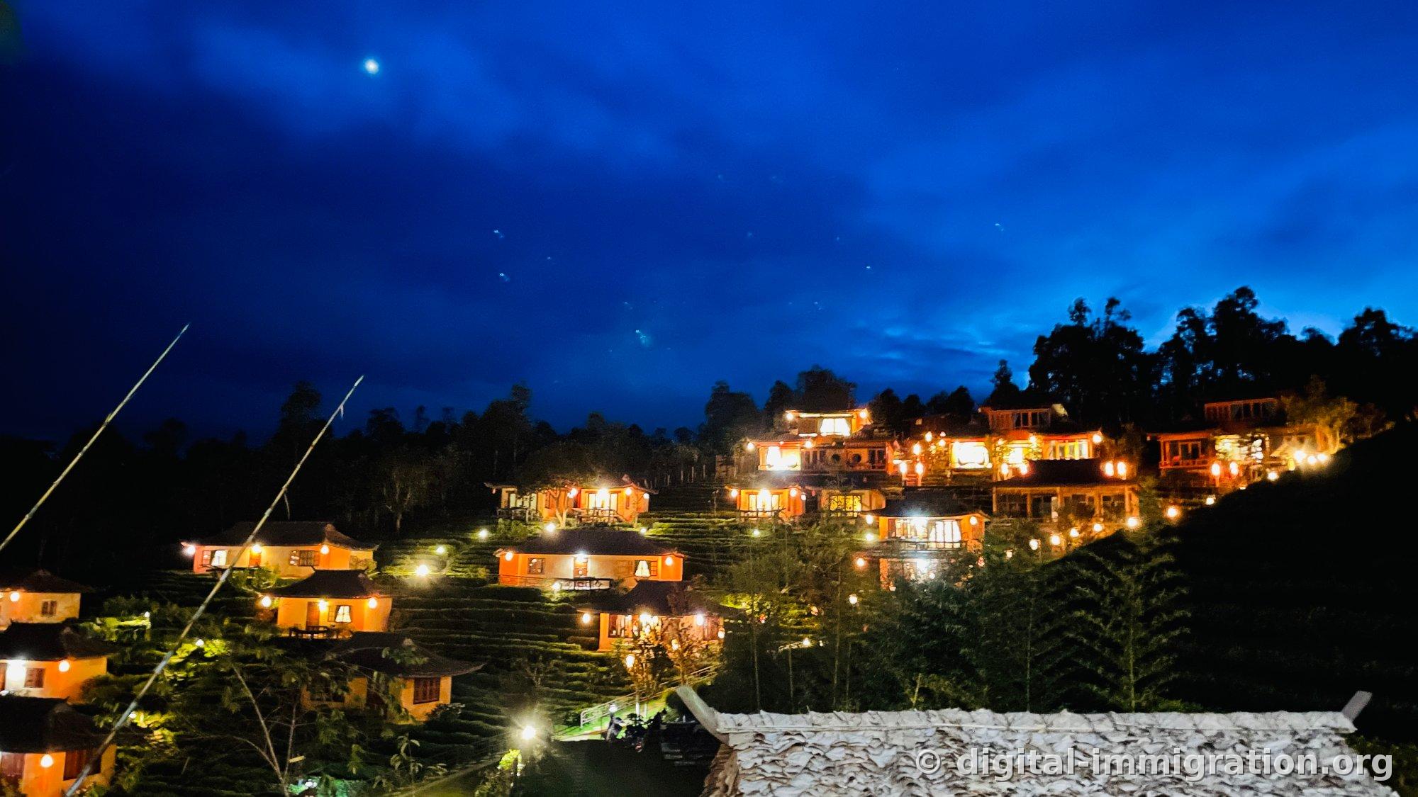 Maisons d'hôtes en terre illuminées par des lumières orange sur la colline de Ban Rak Thai, avec un ciel bleu marine foncé, une lune et des étoiles visibles