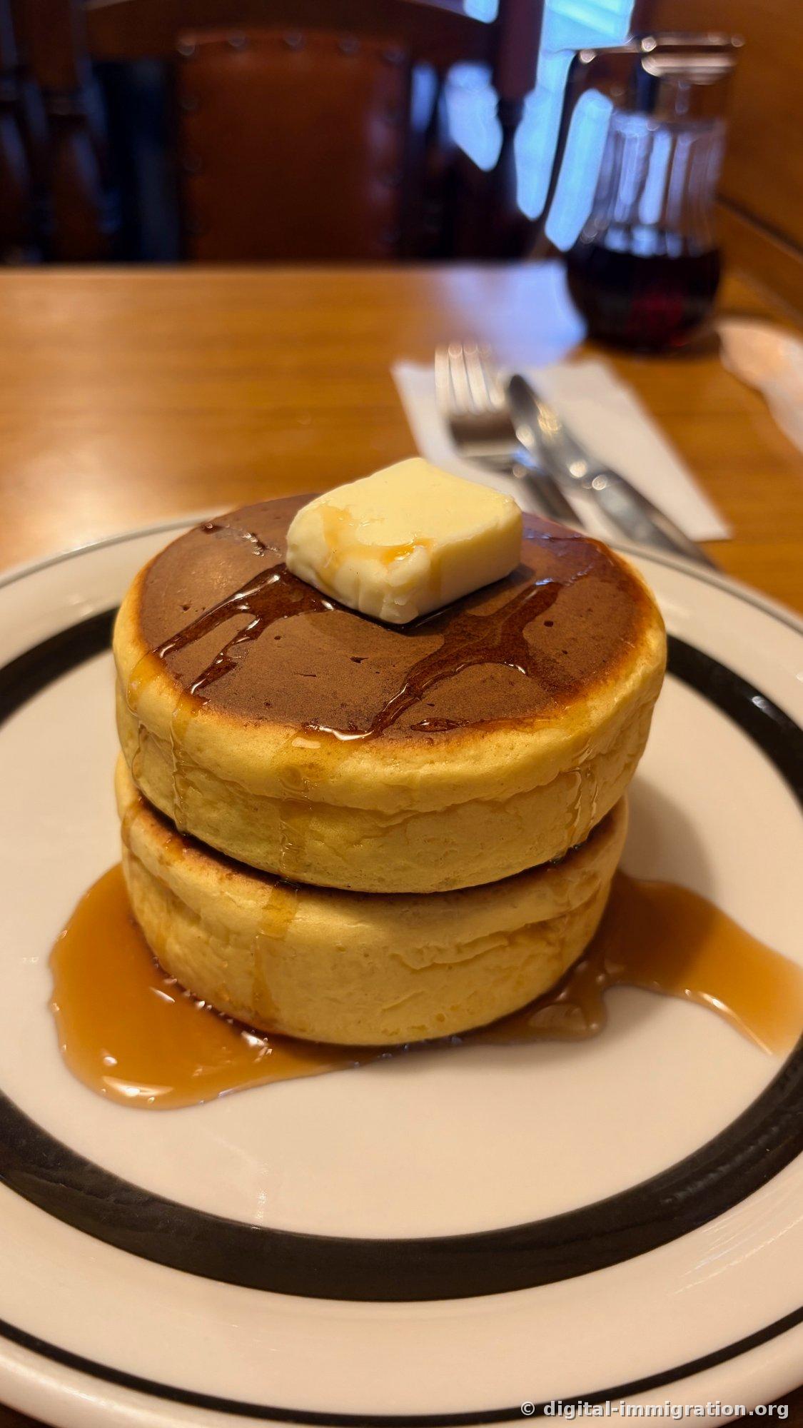 Two thick pancakes stacked on a black-rimmed plate with a rectangular pat of butter on top and syrup streaming down the sides