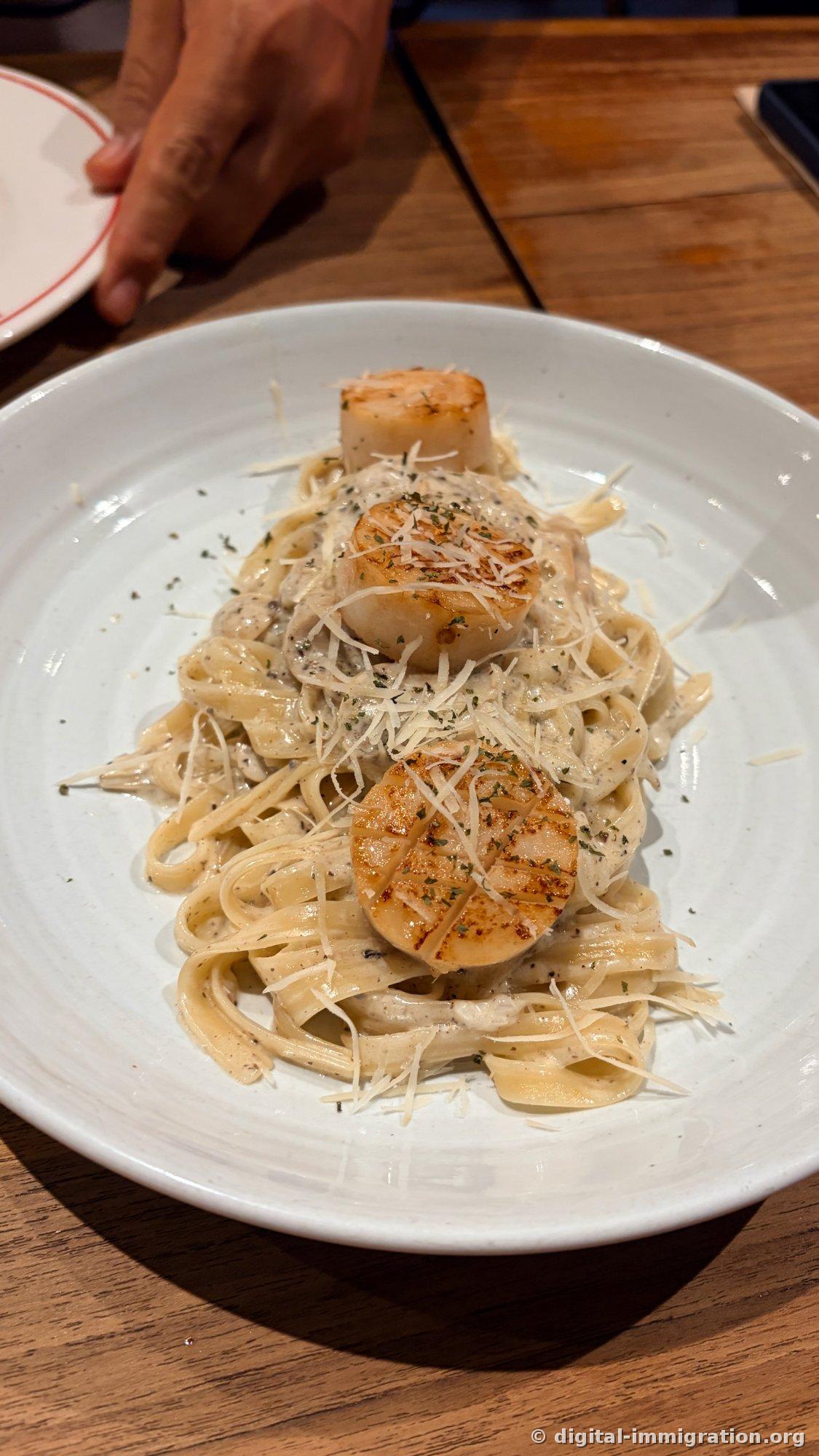 Un assiette blanche de fettuccini à la sauce crème avec trois gros pétoncles dorés à la surface et du parmesan râpé