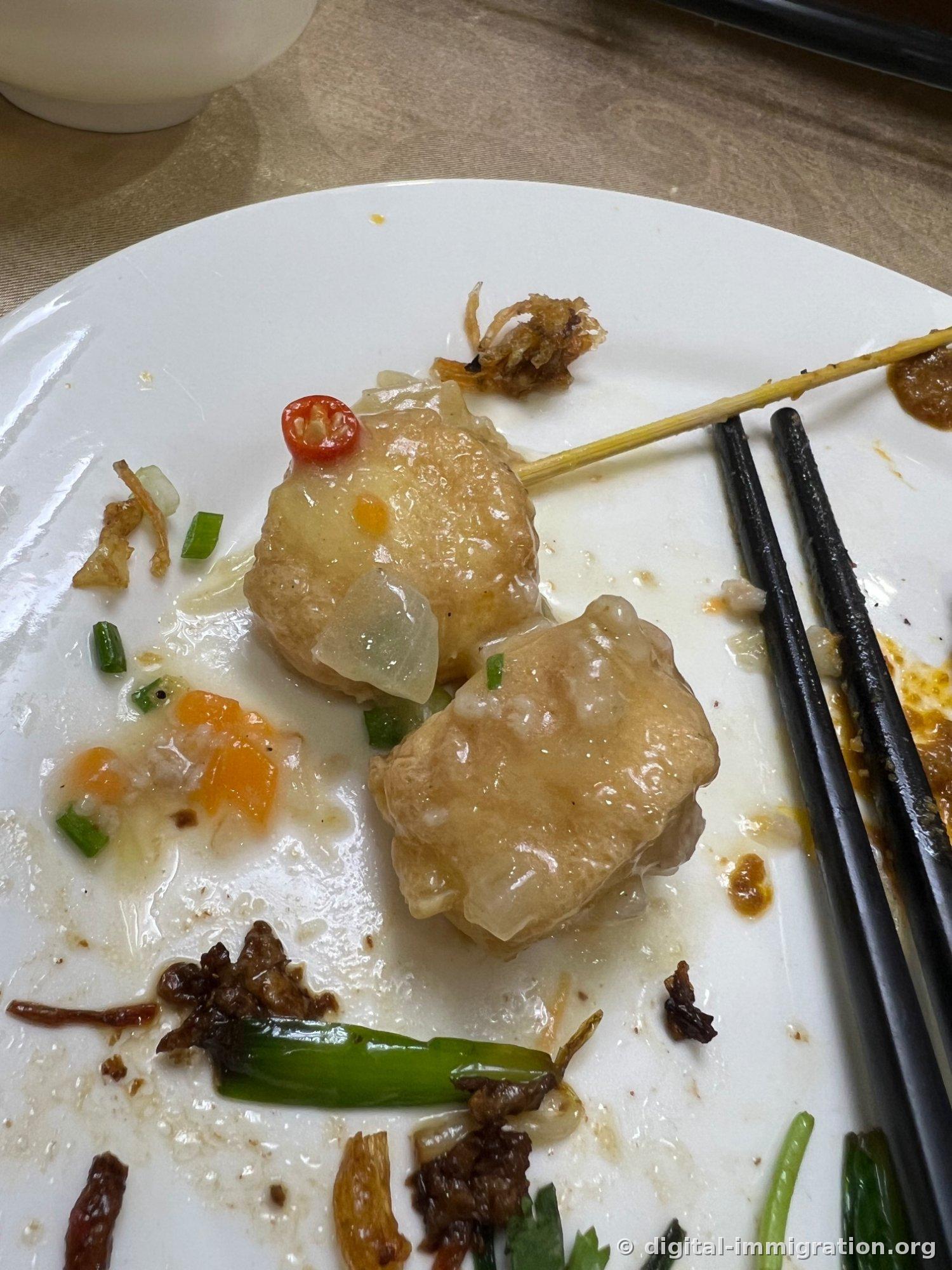 A white plate with two final pieces of fried tofu pockets remaining, drizzled with clear starch sauce, topped with red chili, and surrounded by fried garlic and green onion, with black chopsticks on the side