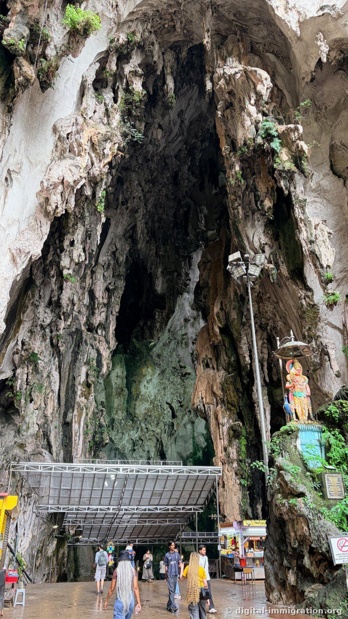 巨大な洞窟のアーチの内部、天井と壁からつらら石が垂れており、下方には灰色の屋根の施設と売店、歩く人々が小さく見える