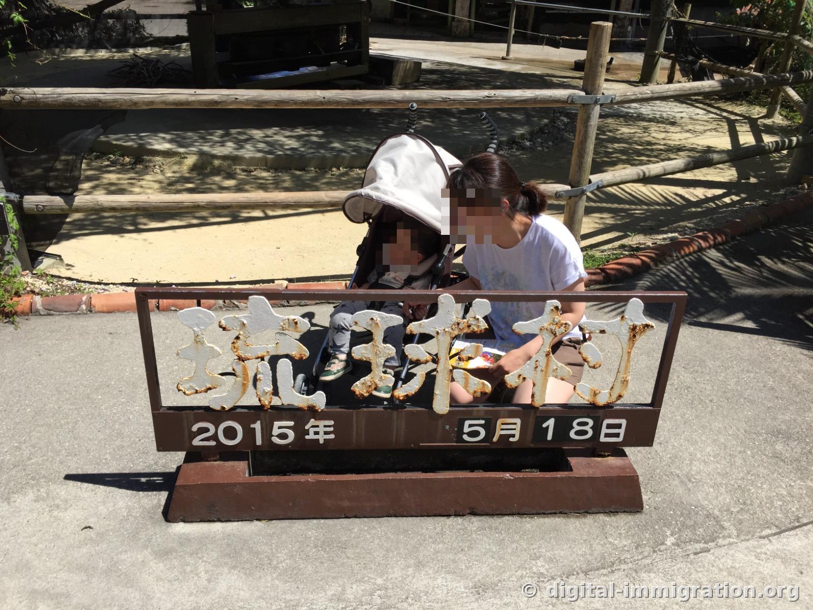 Un panneau de commémoration de date indiquant « 琉球村 2015年 5月 18日 » avec un visiteur assis et une poussette, réalisé derrière le panneau