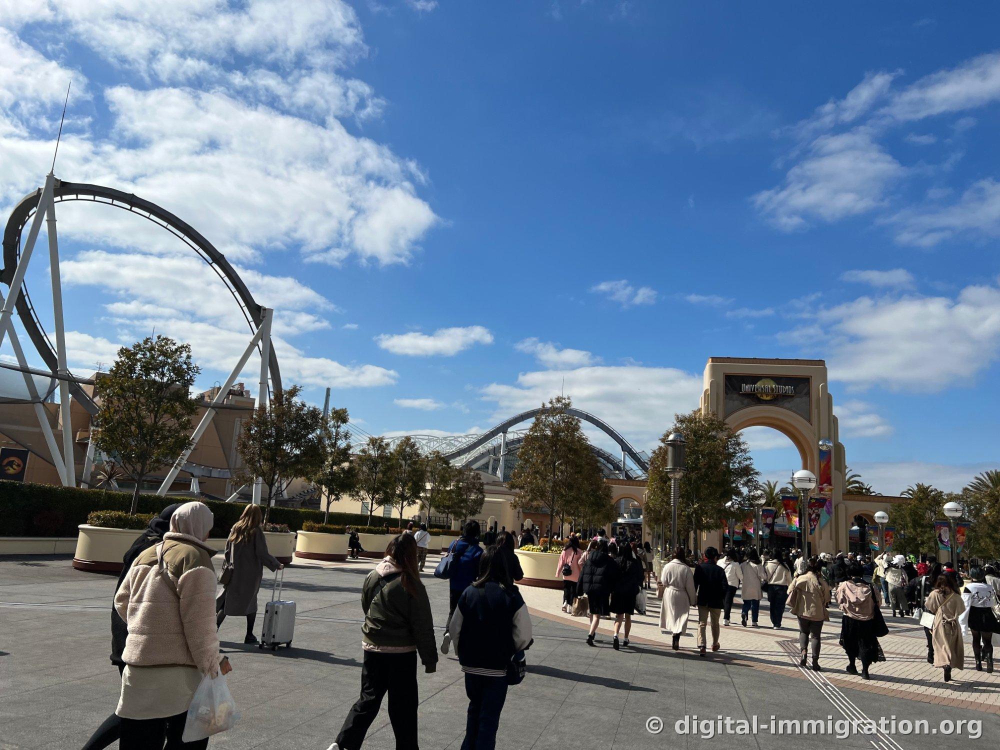 Under a blue sky, visitors walk through Universal Studios' arched main gate while a roller coaster loop structure appears on the left side of the plaza