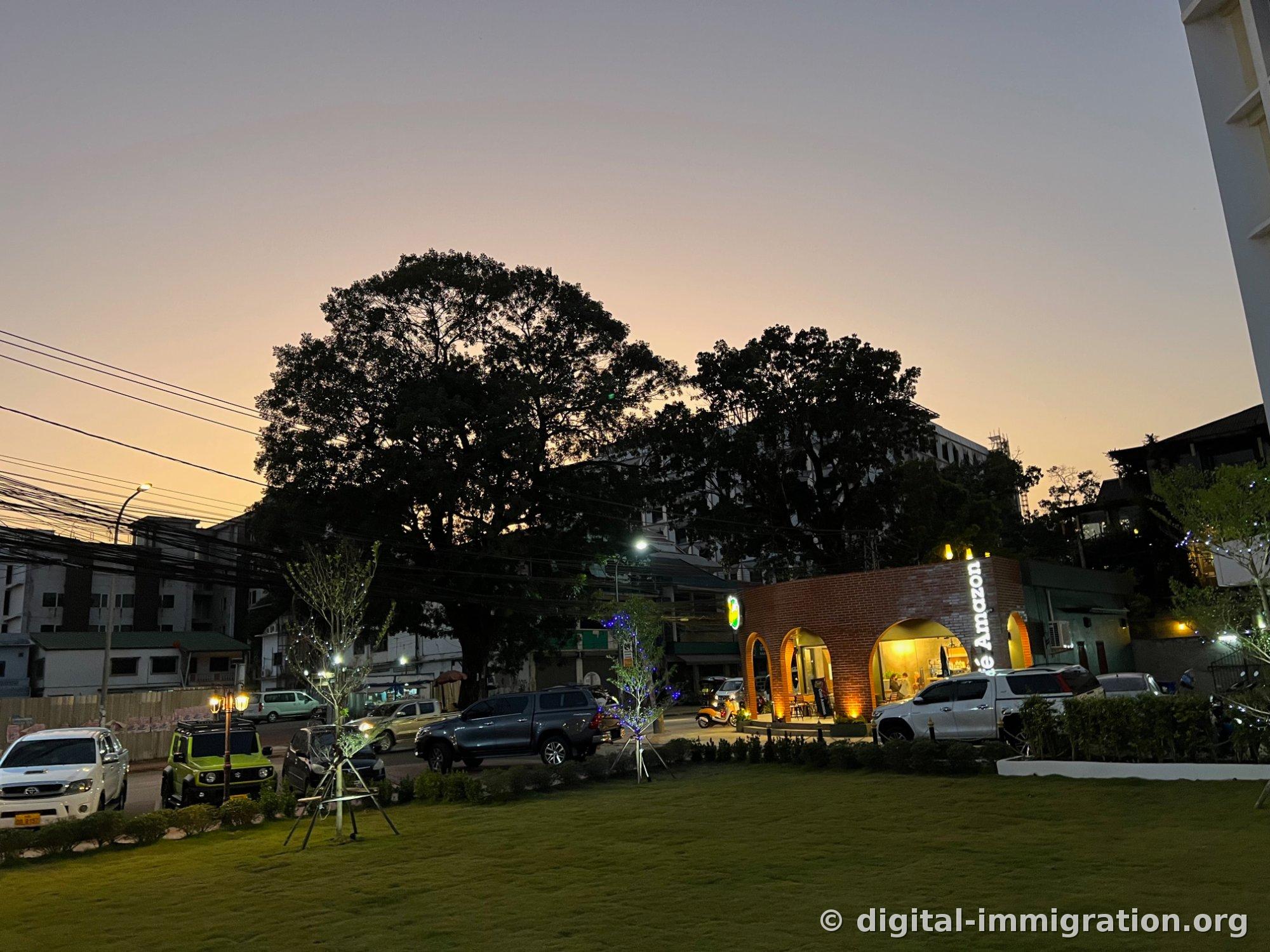 Le coucher du soleil orange avec la silhouette d'un grand arbre et le bâtiment en brique aux arches du Café Amazon, avec pelouse et camionnettes garées devant
