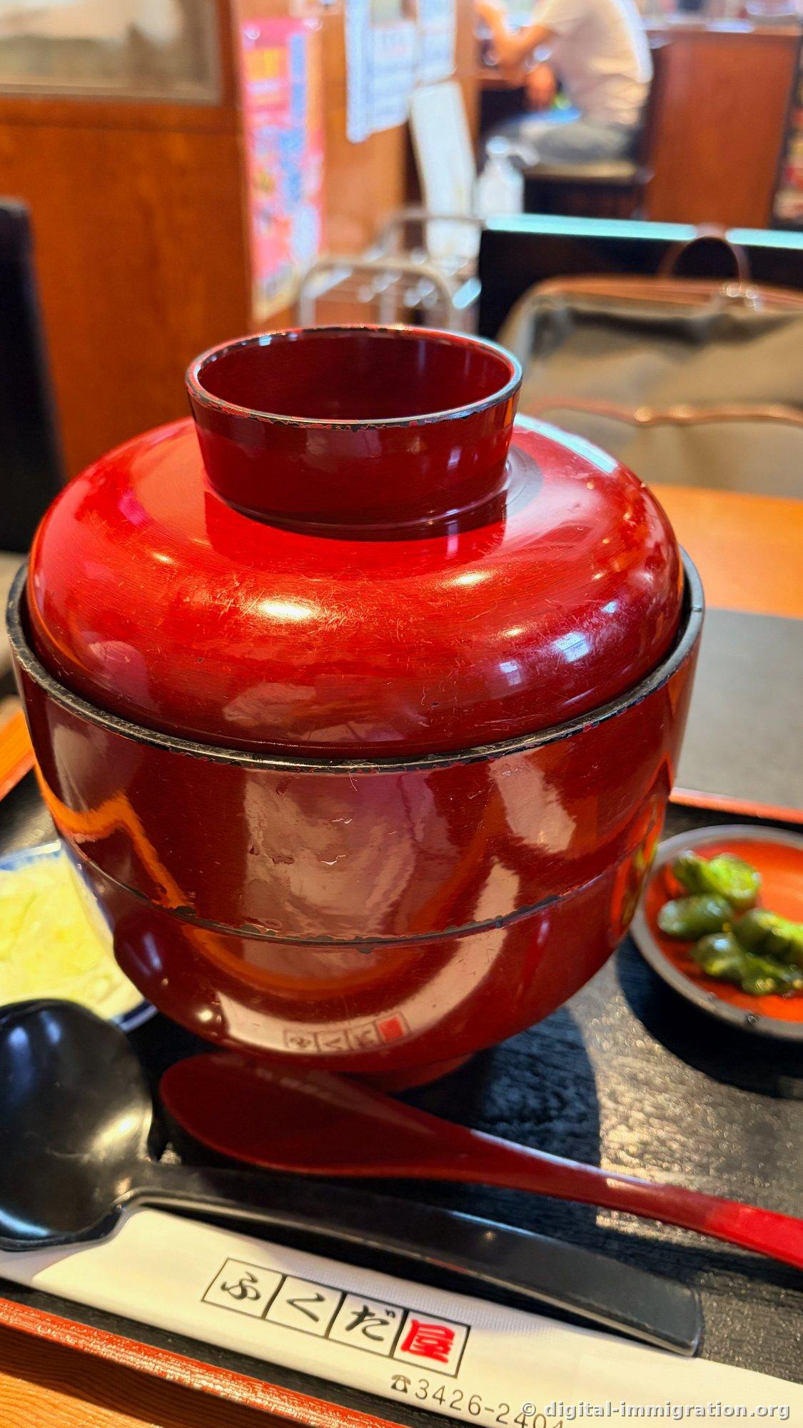 Bol donburi recouvert d'un couvercle vernis rouge, condiment mariné vert à côté, cuillère noire, emballage de baguettes imprimé avec 'ふくだ屋' et le numéro de téléphone