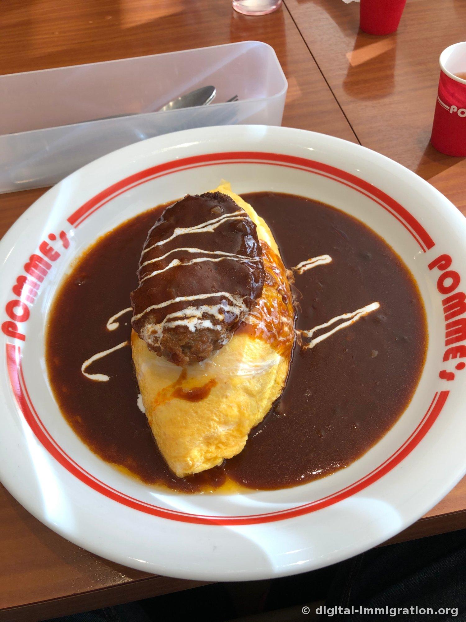 Vue d'ensemble du plat : une assiette blanche avec le logo POMME's gravé, recouverte d'une sauce demi-glace foncée, l'omurice jaune dessus, et un steak haché garni de sauce et de traits blancs de crème