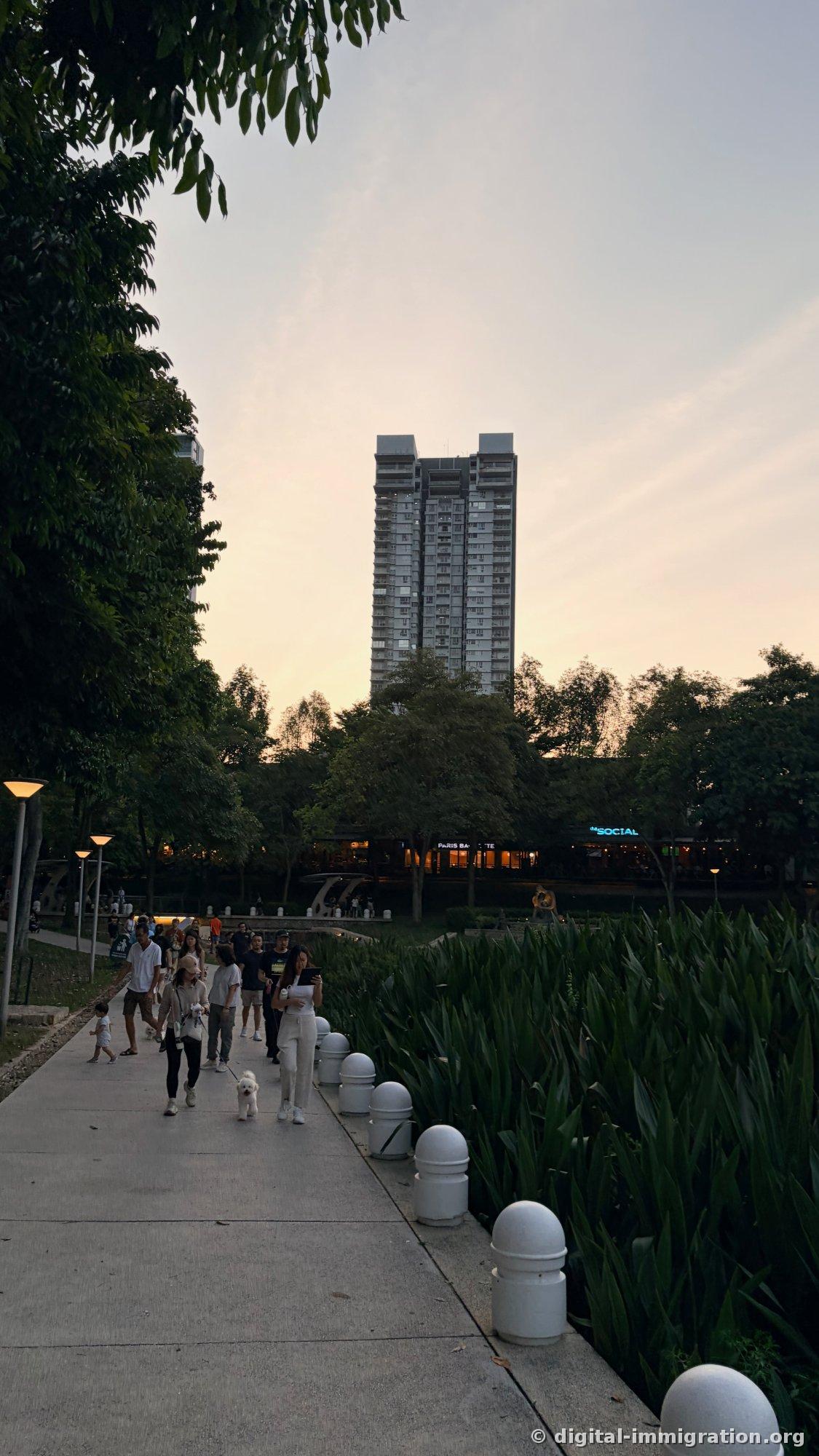 해질녘 산책로를 걸어오는 사람들과 흰 비숑, 호수 건너편에 the SOCIAL 파란 네온 간판과 PARIS BAGUETTE 매장 불빛, 뒤로 고층 콘도 실루엣