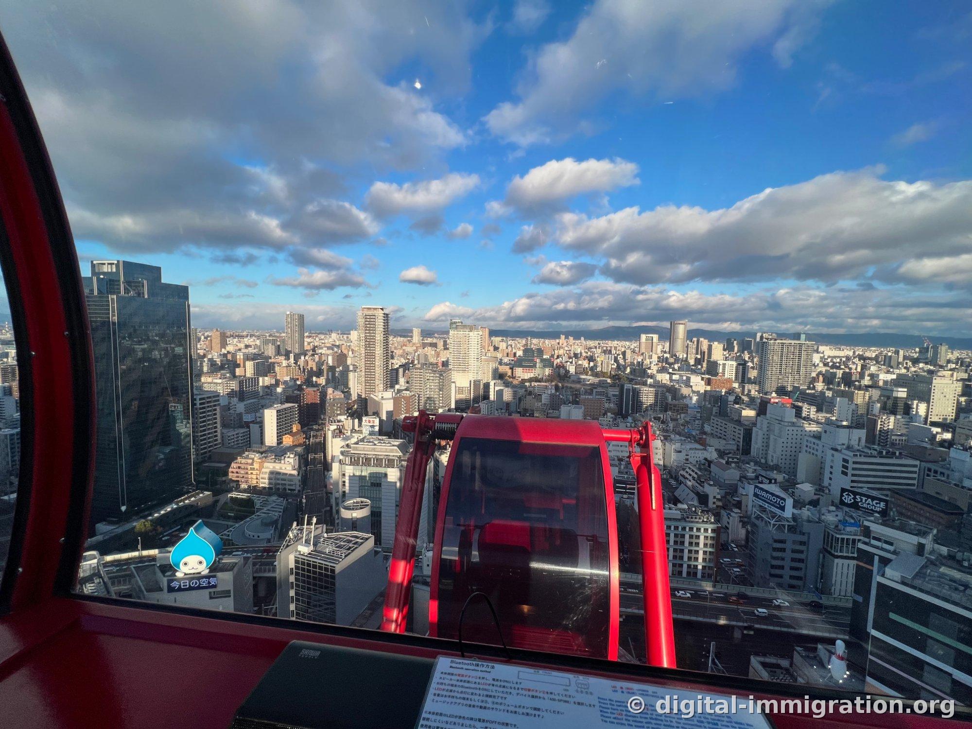 カプセルの窓越しに、先行する赤いカプセルと大阪市街地、座席前に置かれたBluetoothコントロールガイドが見える室内視点