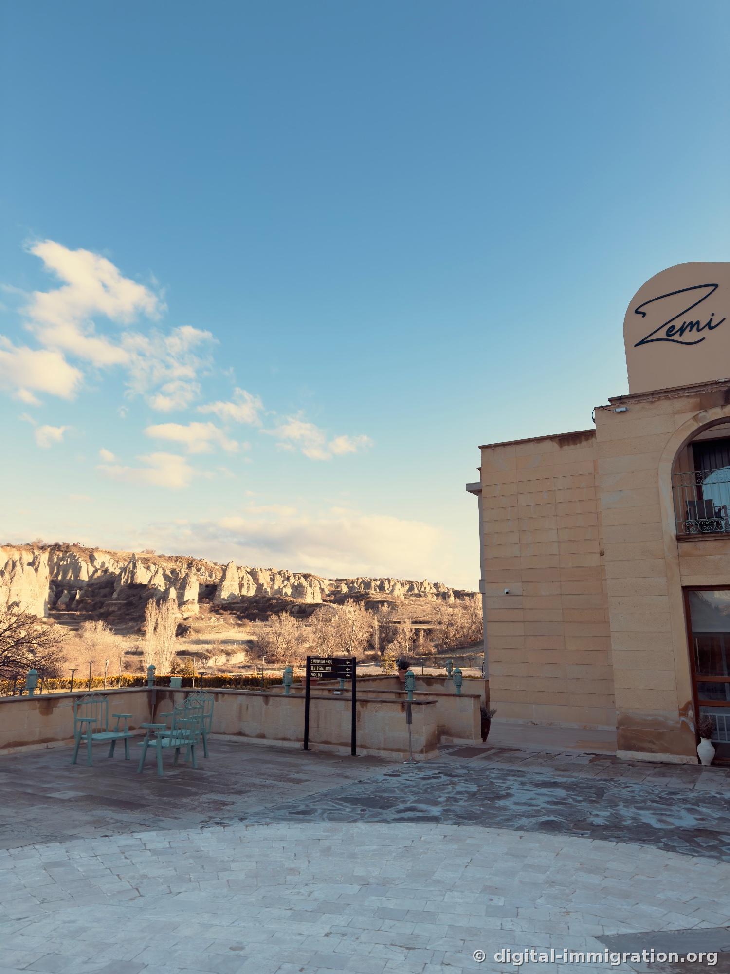 Depuis la terrasse de l'hôtel, la crête des cheminées de fée et des chaises métalliques menthe, un panneau de direction 'SWIMMING POOL / ZEMI RESTAURANT / POOL BAR'