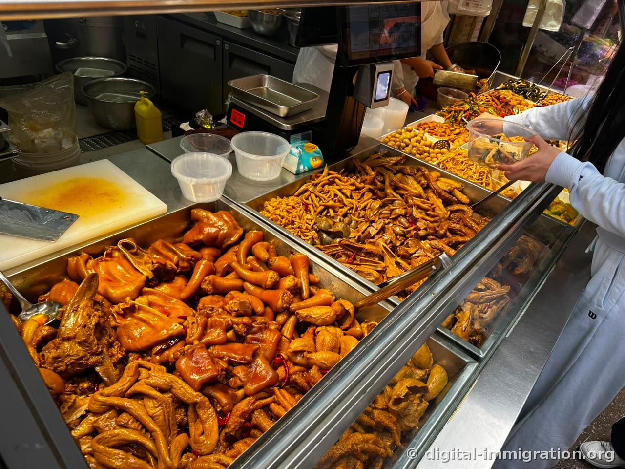 A glass display case with compartments of braised pig trotters and ears, chicken feet, duck necks, braised eggs and seasoned vegetables, with a scale and customer's hands placing items in a container