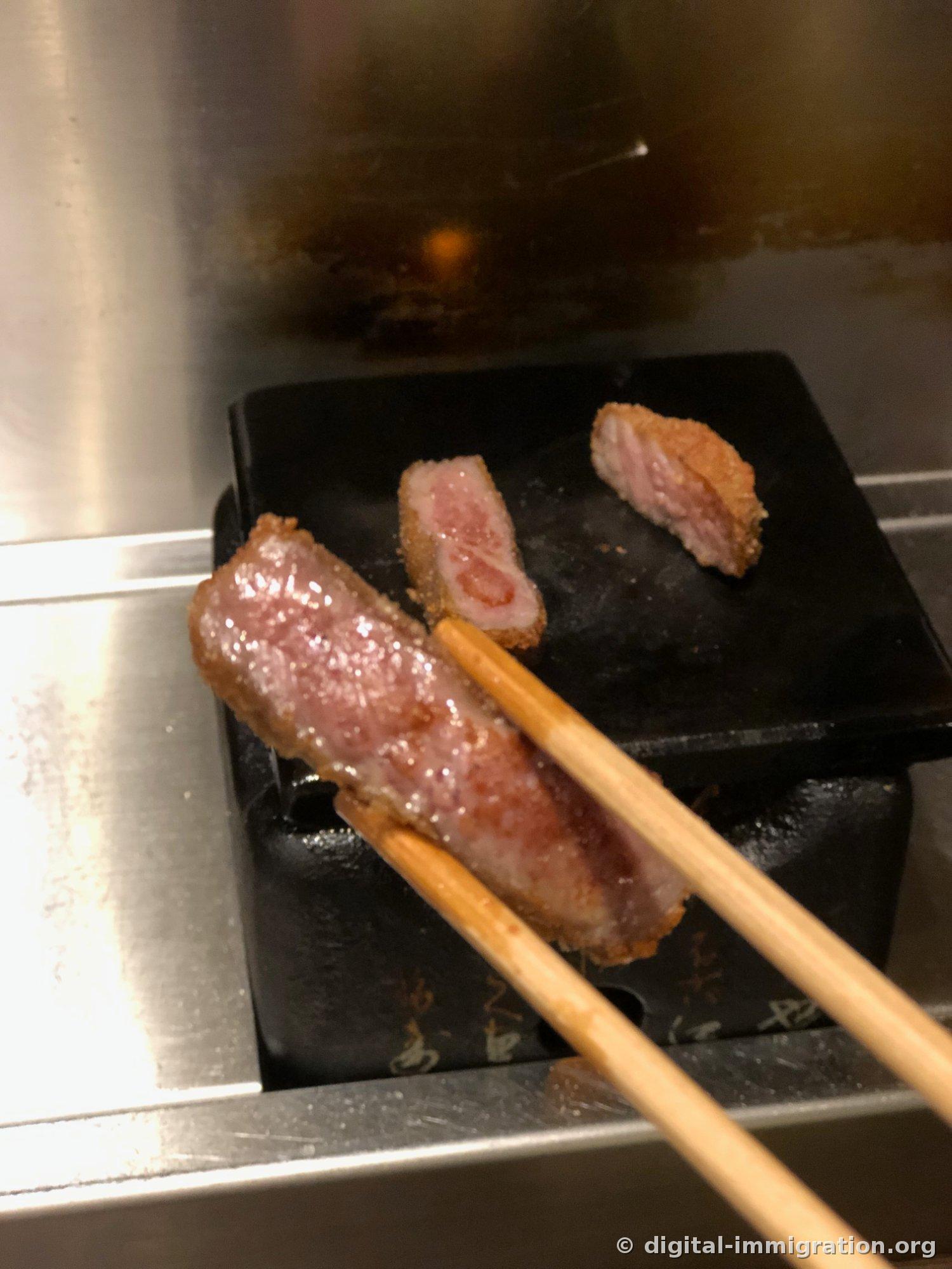 Un morceau de Gyukatsu cuit saisi avec des baguettes en bois. Deux morceaux restent encore sur la pierre