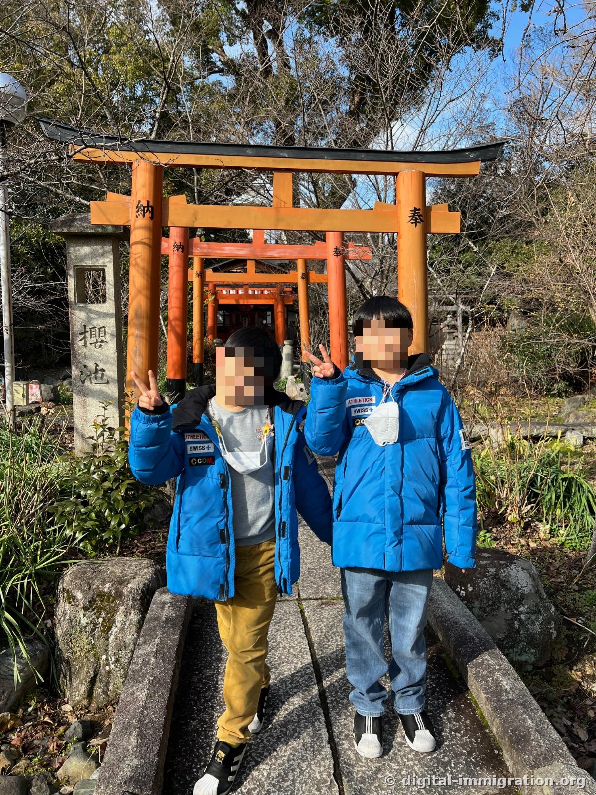 A narrow path lined with small orange torii gates marked '奉納', beside a stone pillar inscribed with '櫻池', where two children wearing blue padded jackets make a peace sign pose